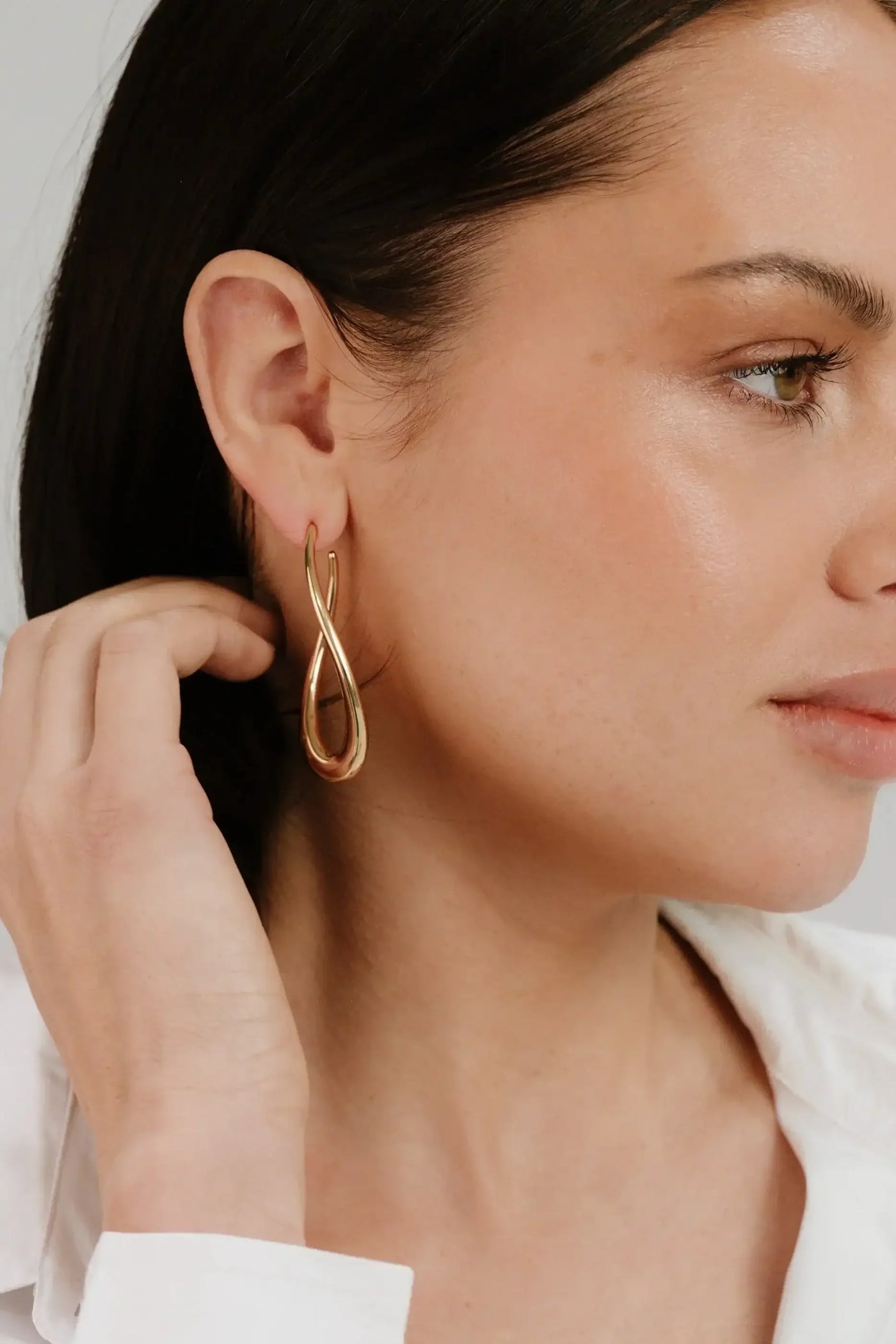 A woman is gently touching her ear while wearing a stylish, elongated gold earring. She has shoulder-length hair and appears to be in a neutral-colored setting.