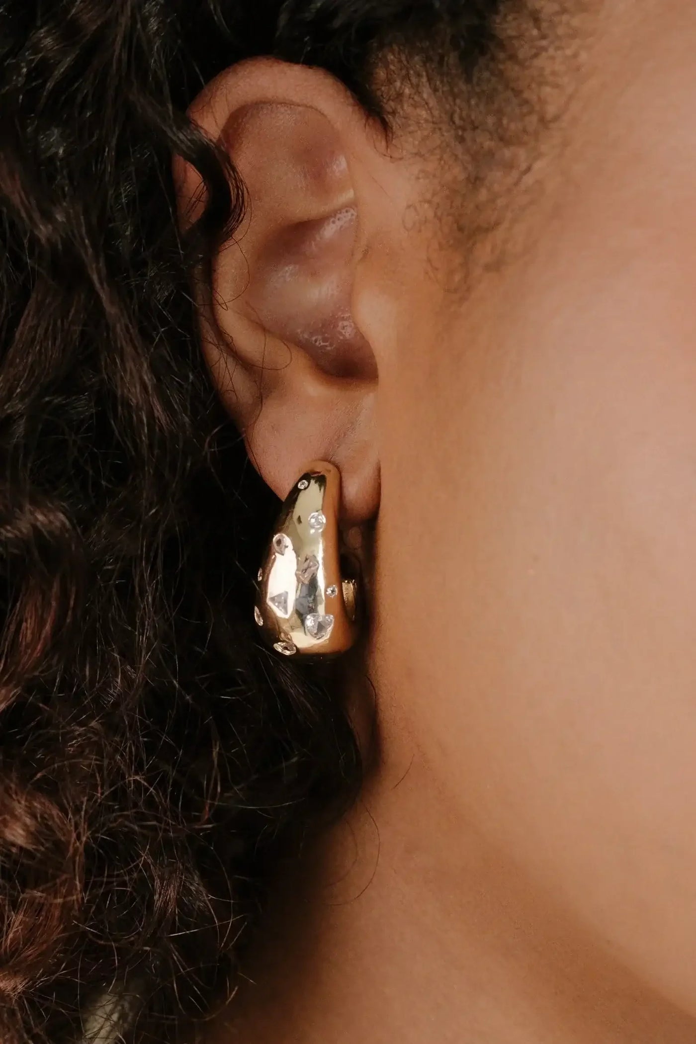 A gold earring with a shiny, textured surface and small decorative stones adorns the lobe of a person's ear. They have curly hair framing their face.