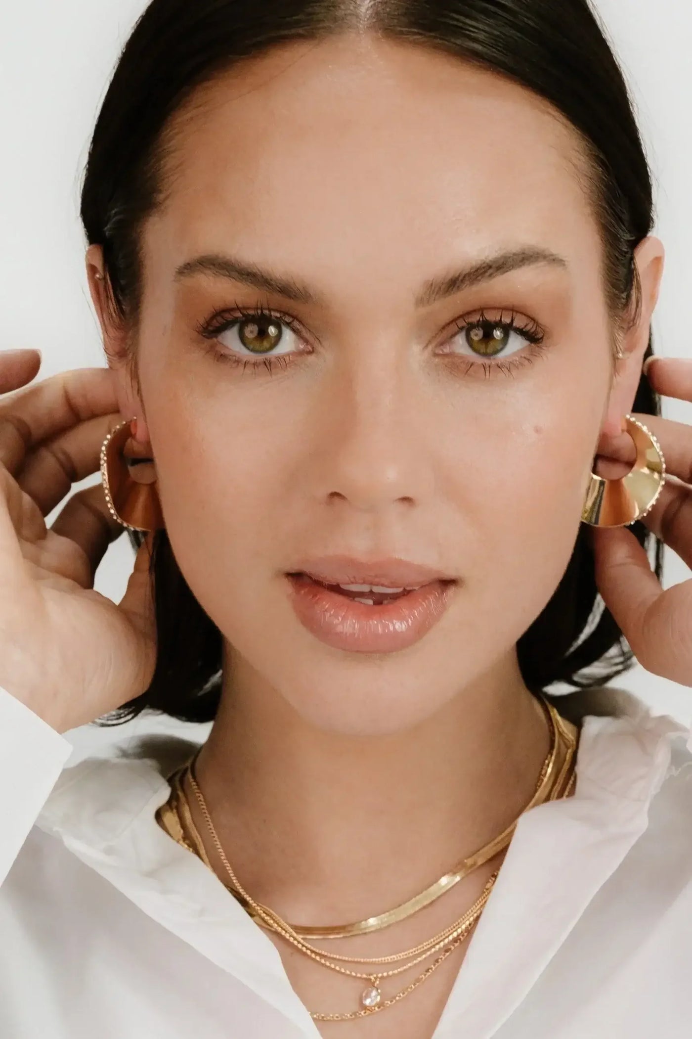 A woman with long, dark hair is adjusting large hoop earrings with both hands. She wears layered gold necklaces and a white shirt, in a well-lit, neutral background.