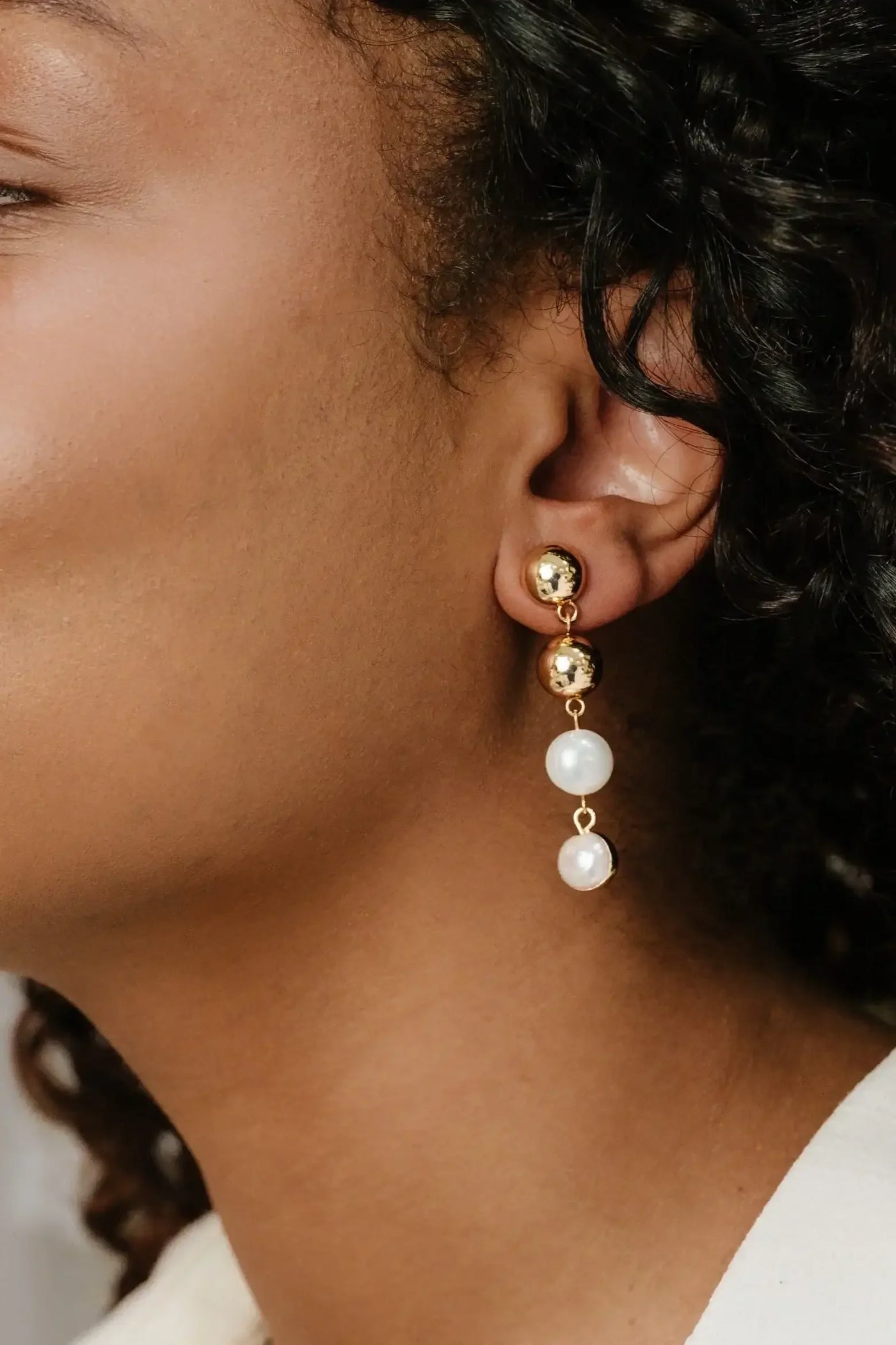 A close-up view of a woman's ear showcases a gold and pearl earring. The earring features two shiny gold spheres and two dangling white pearls, highlighting a stylish accessory against smooth skin.