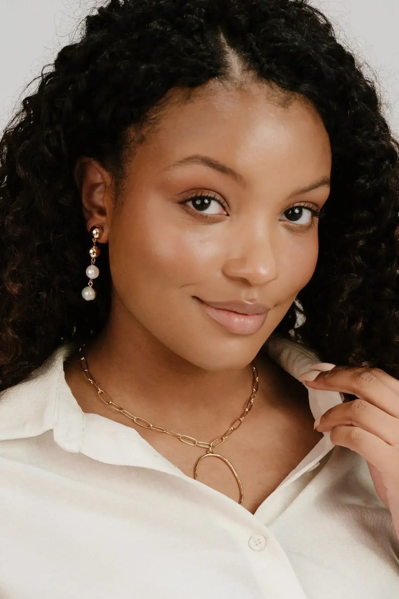 A young woman with curly hair is smiling while touching her collar. She wears a white button-up shirt and a gold necklace with a circular pendant, showcasing elegant drop earrings.