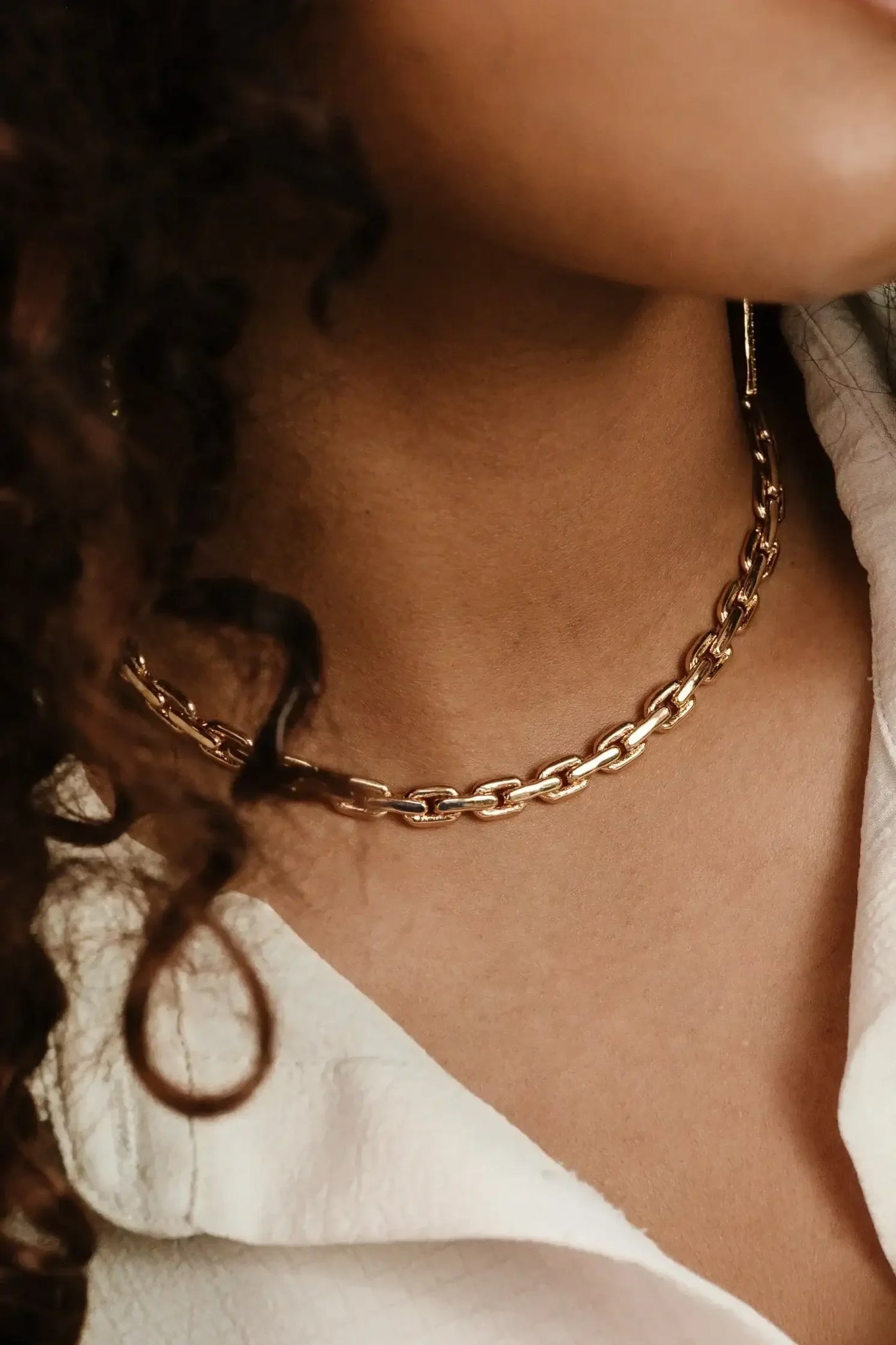 A gold chain necklace rests against the skin of a person's neck, with loose curls of hair partially visible. The setting suggests a casual, stylish environment.
