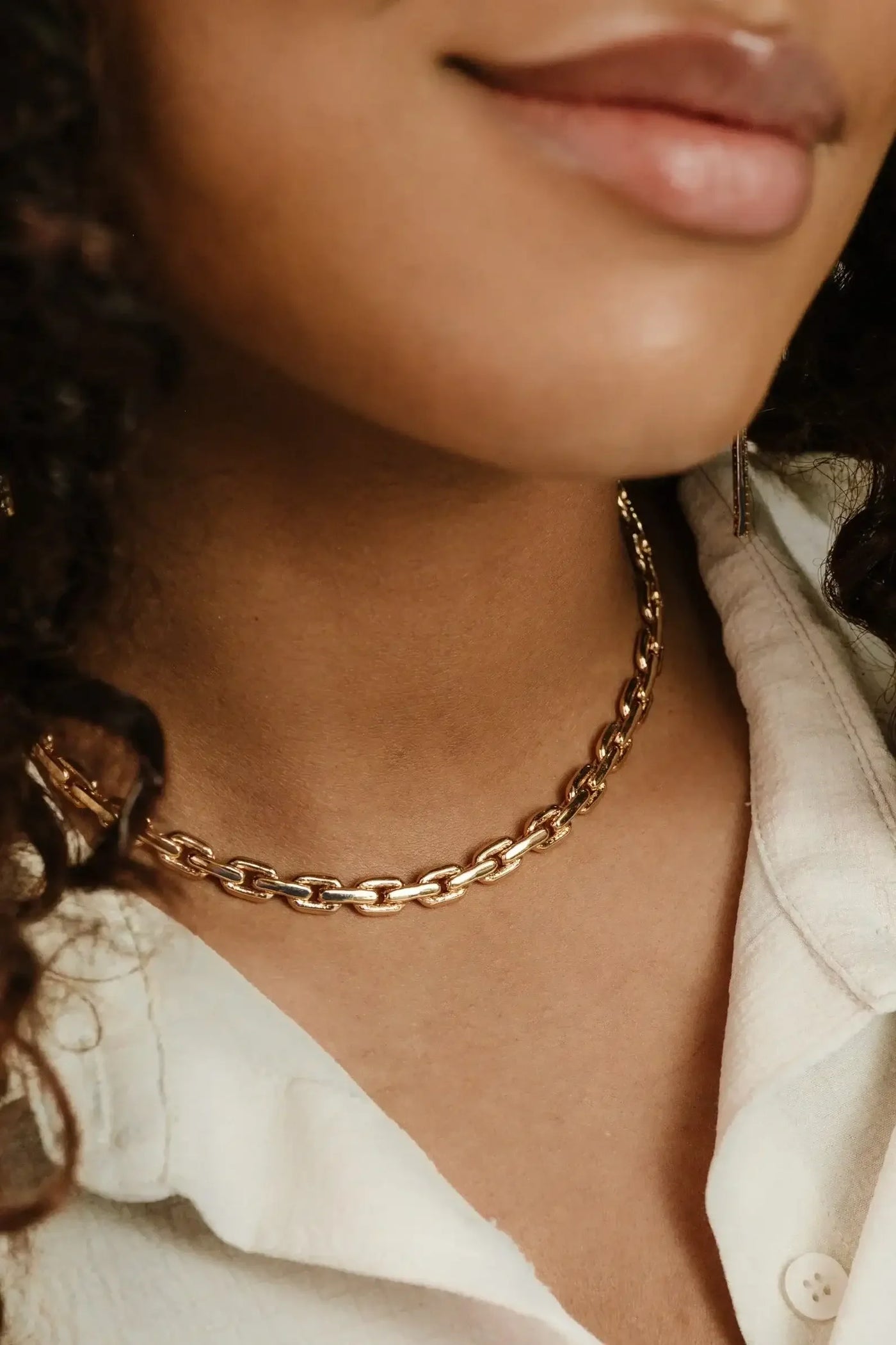 A gold chain necklace rests around a neck, complementing smooth skin and soft curls. The individual wears a light-colored, buttoned shirt, enhancing the elegant appearance of the jewelry.