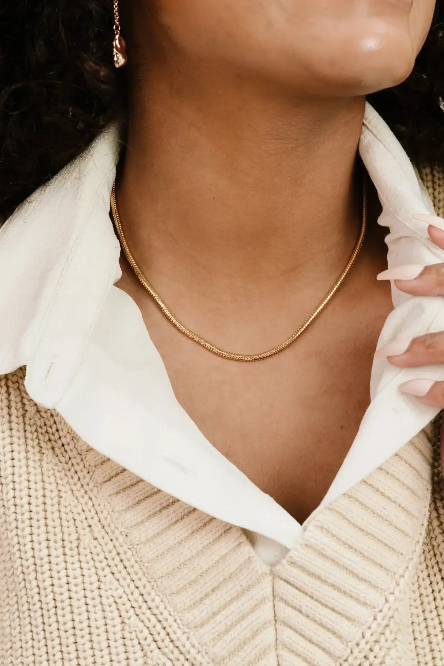 A gold chain necklace rests on a person's neck while they lightly hold their collar, which features a white shirt under a beige knitted sweater. The setting suggests a casual, relaxed atmosphere.