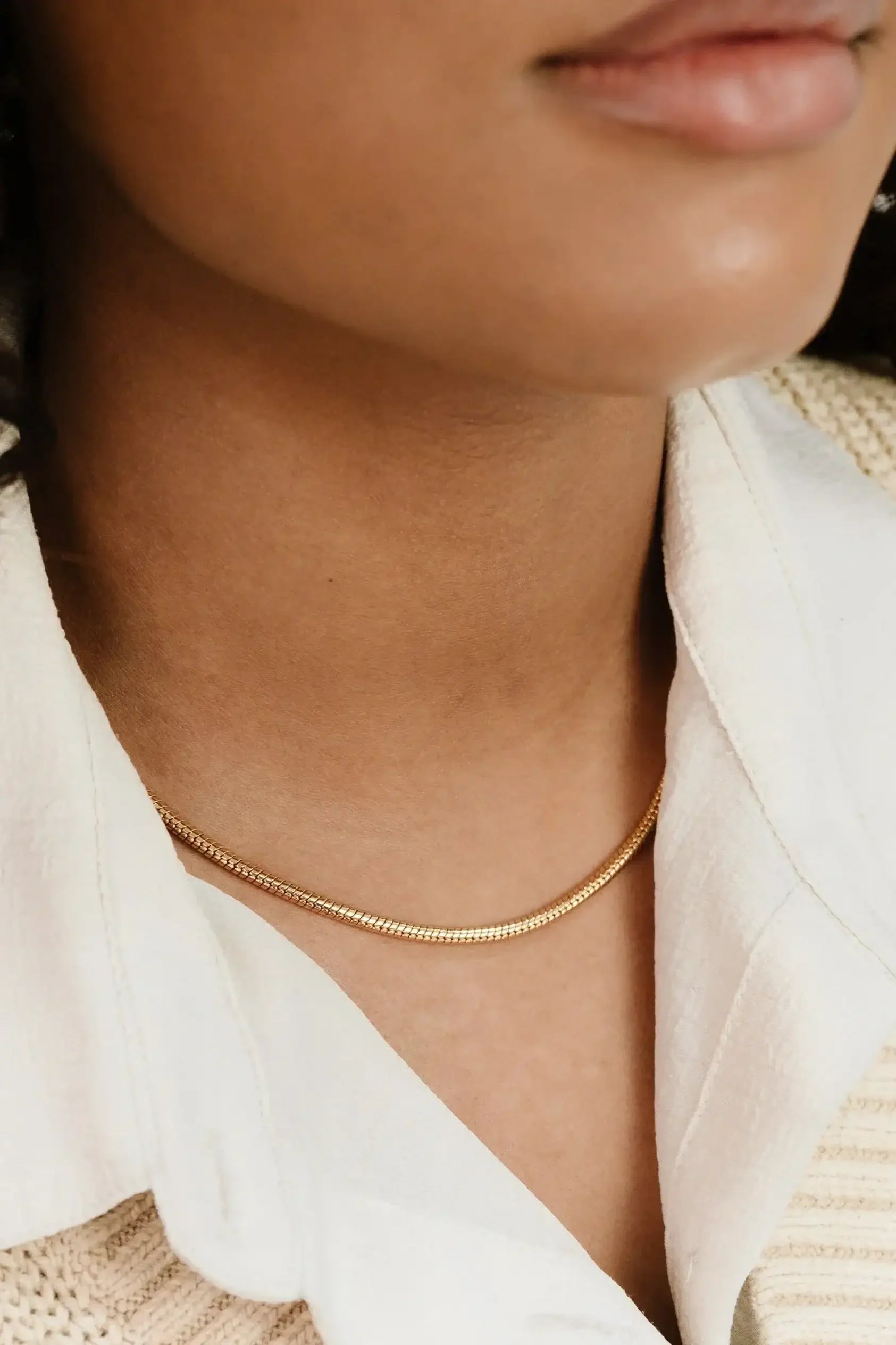 A close-up of a person's neck features a shiny gold necklace resting against smooth skin. The collar of a white shirt is partially visible, suggesting a casual but elegant style.