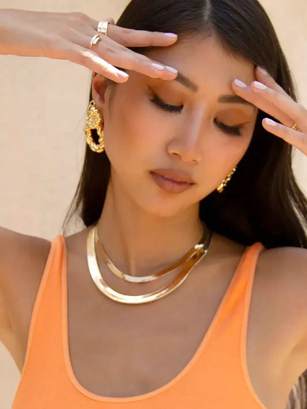 A woman wears a vibrant orange tank top and gold jewelry, gently touching her forehead with closed eyes, set against a light, neutral background that enhances her serene expression.