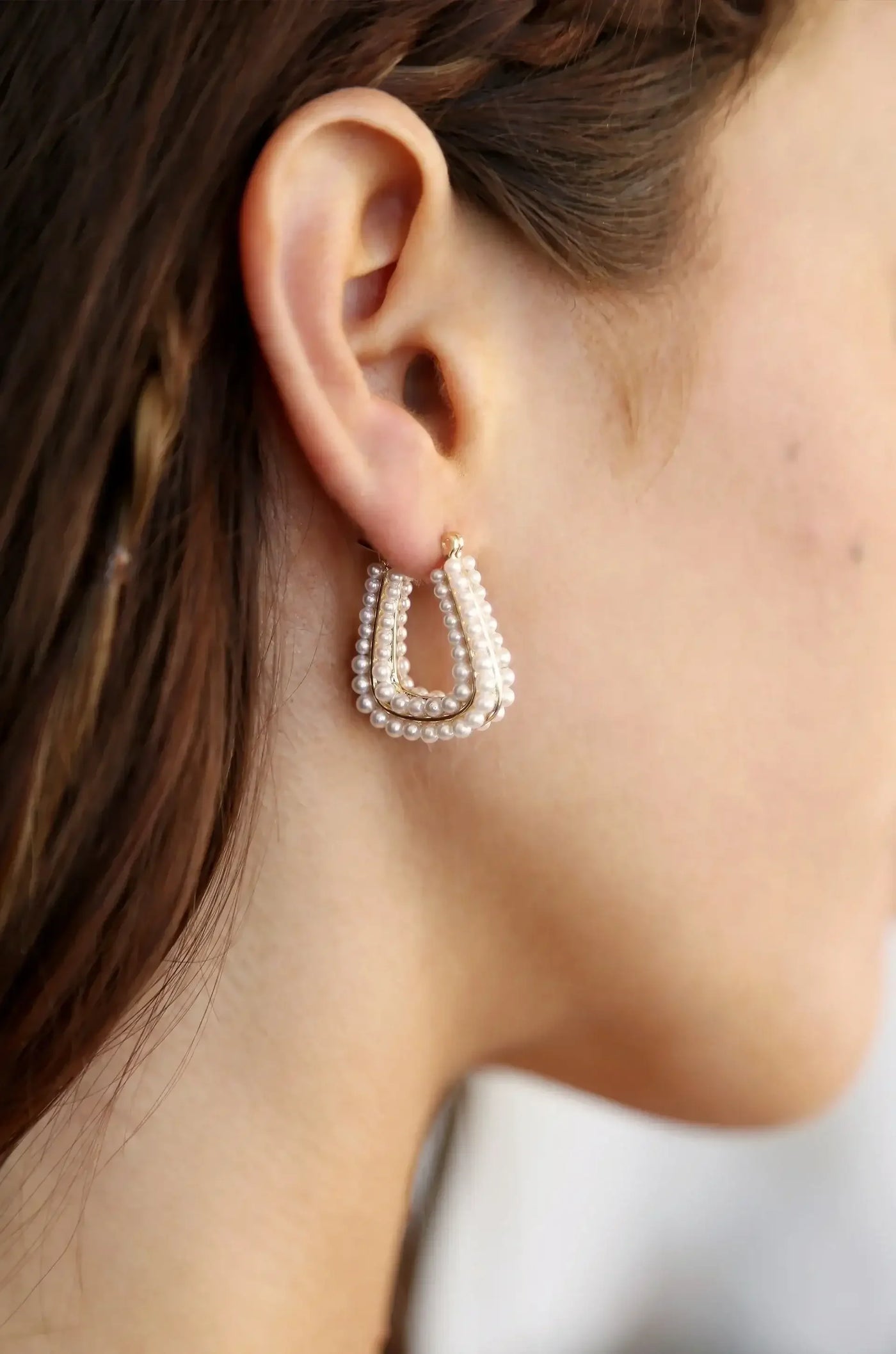 A decorative earring hangs from a woman's ear. It features a gold outline adorned with white pearls, accentuating the elegant curve of her neck against a softly blurred background.