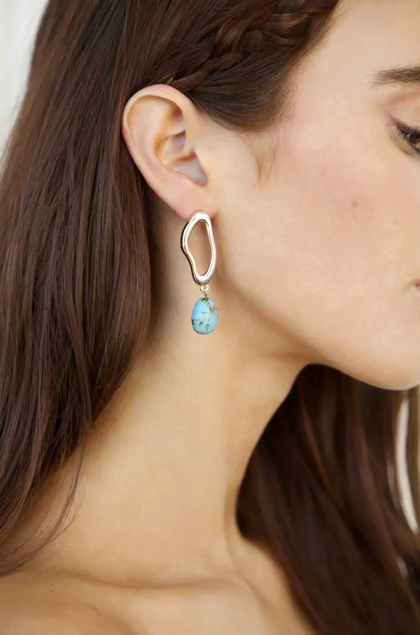 A large, abstract gold earring hangs from a woman's ear, featuring a turquoise stone beneath it. The setting is neutral and soft, highlighting her side profile.