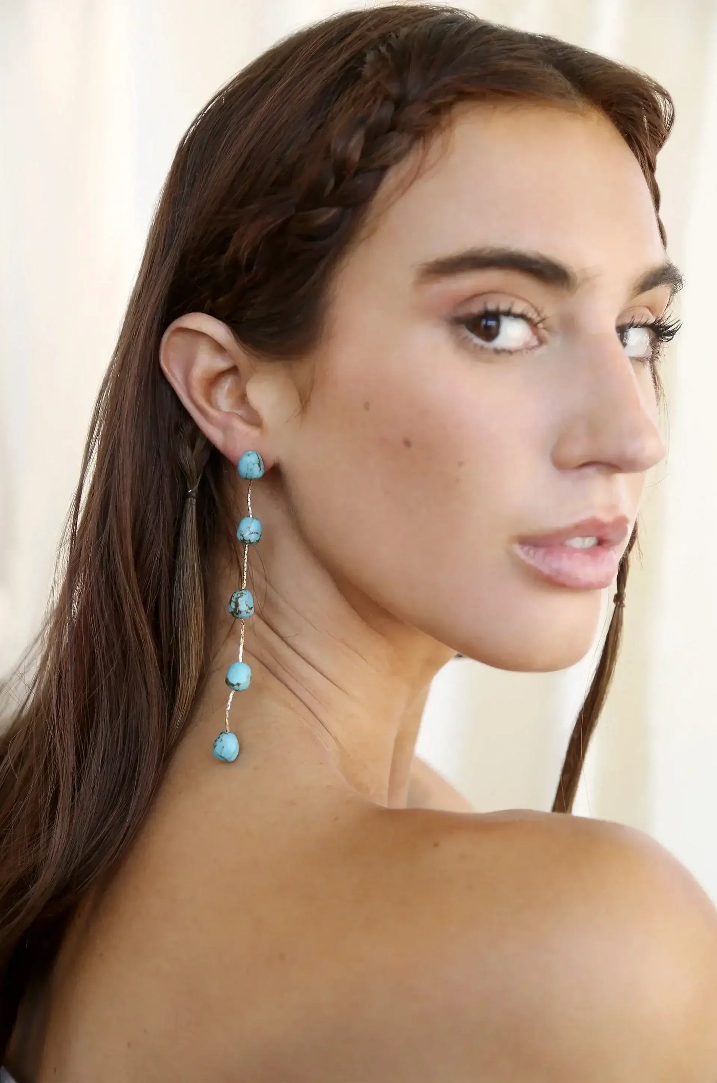 A woman with long, brown hair featuring a braid poses, showcasing a pair of dangling turquoise earrings against a soft, light-colored background. Her expression is serene and thoughtful.