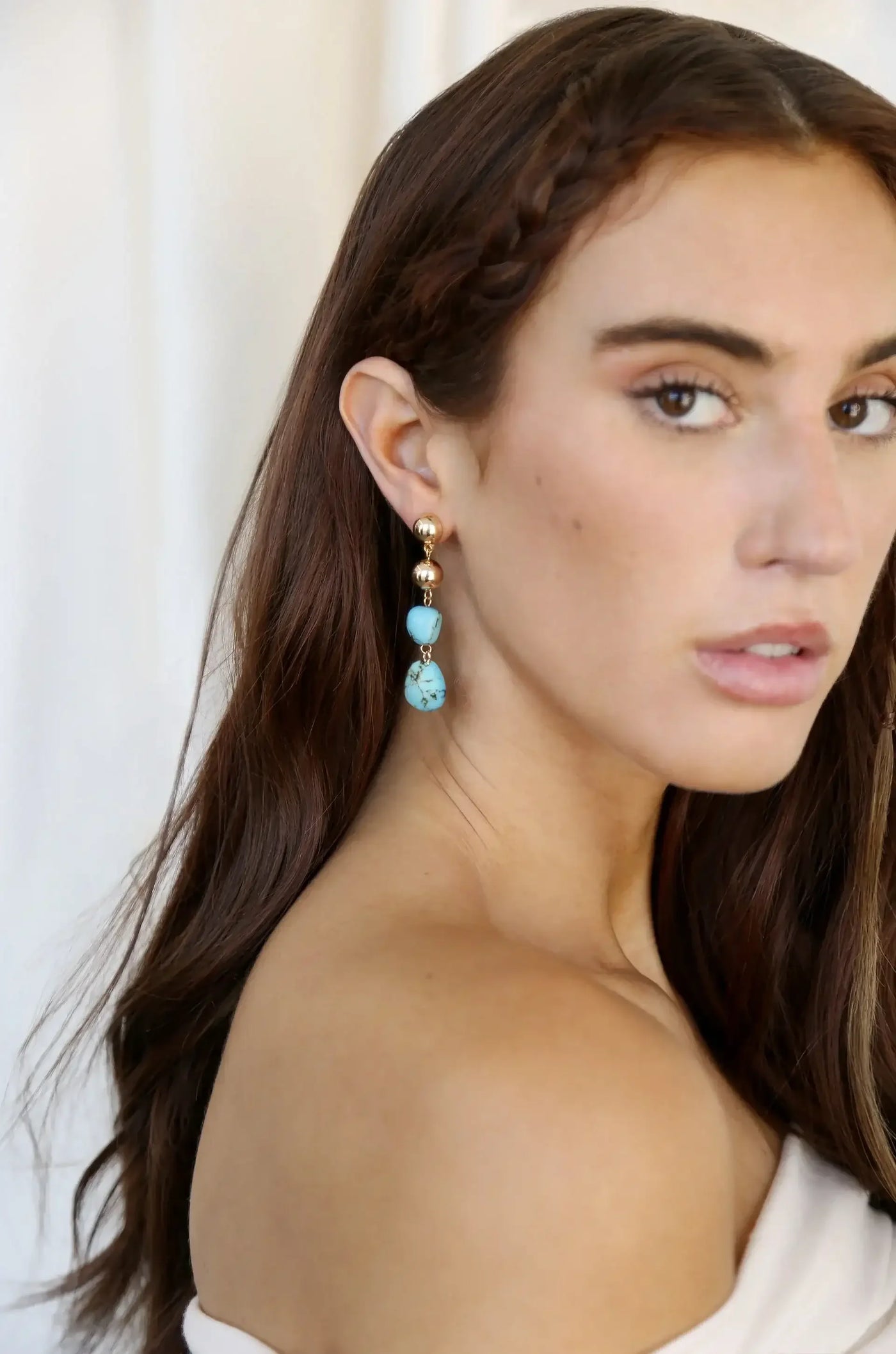 A young woman with long, brown hair wears elegant, dangling earrings featuring gold and turquoise elements. She gazes directly at the viewer against a soft, neutral background.