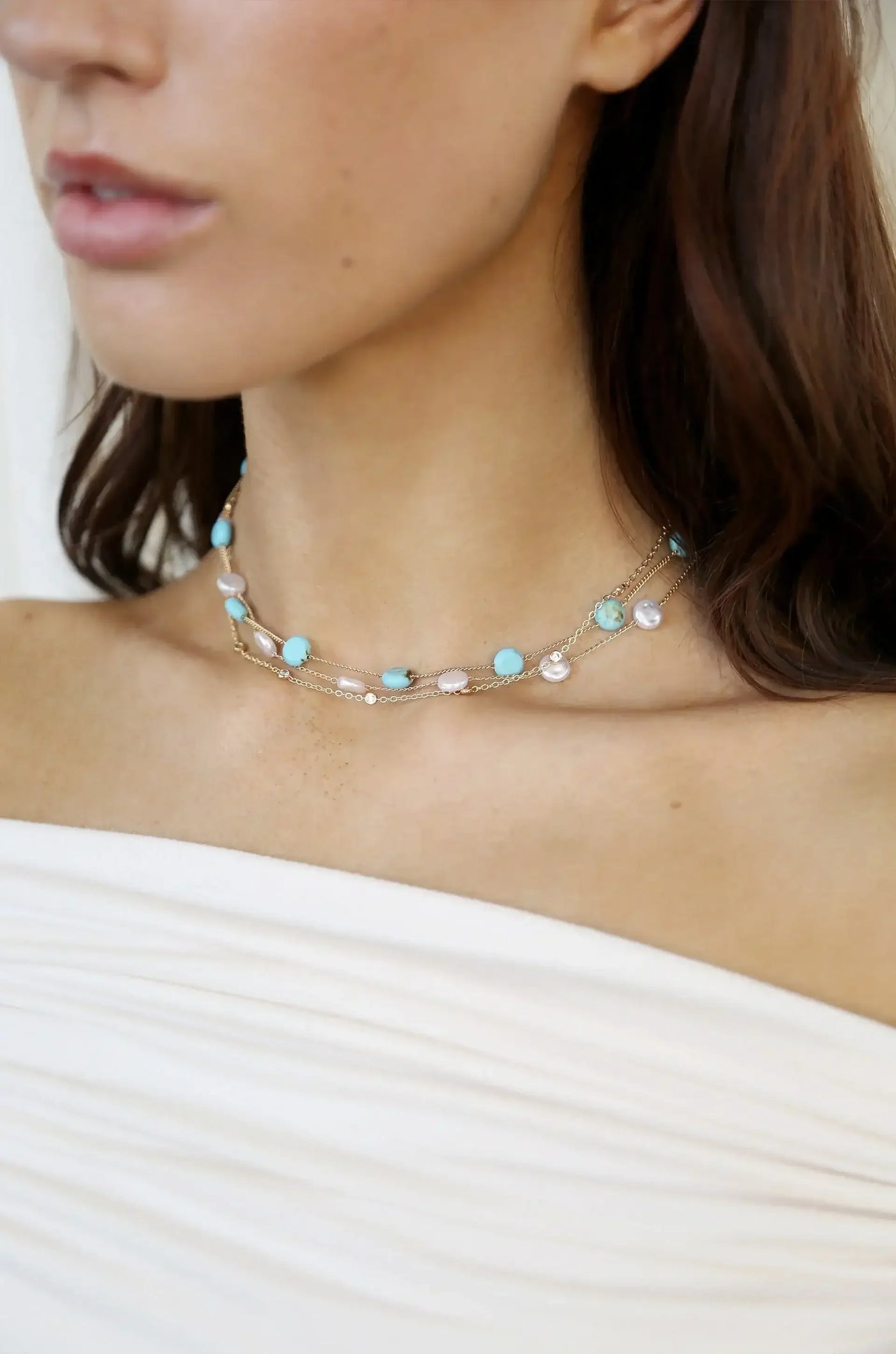 A delicate necklace featuring turquoise and pearl beads adorns a person's neck, complementing their bare shoulders dressed in soft, white fabric, set against a light background.