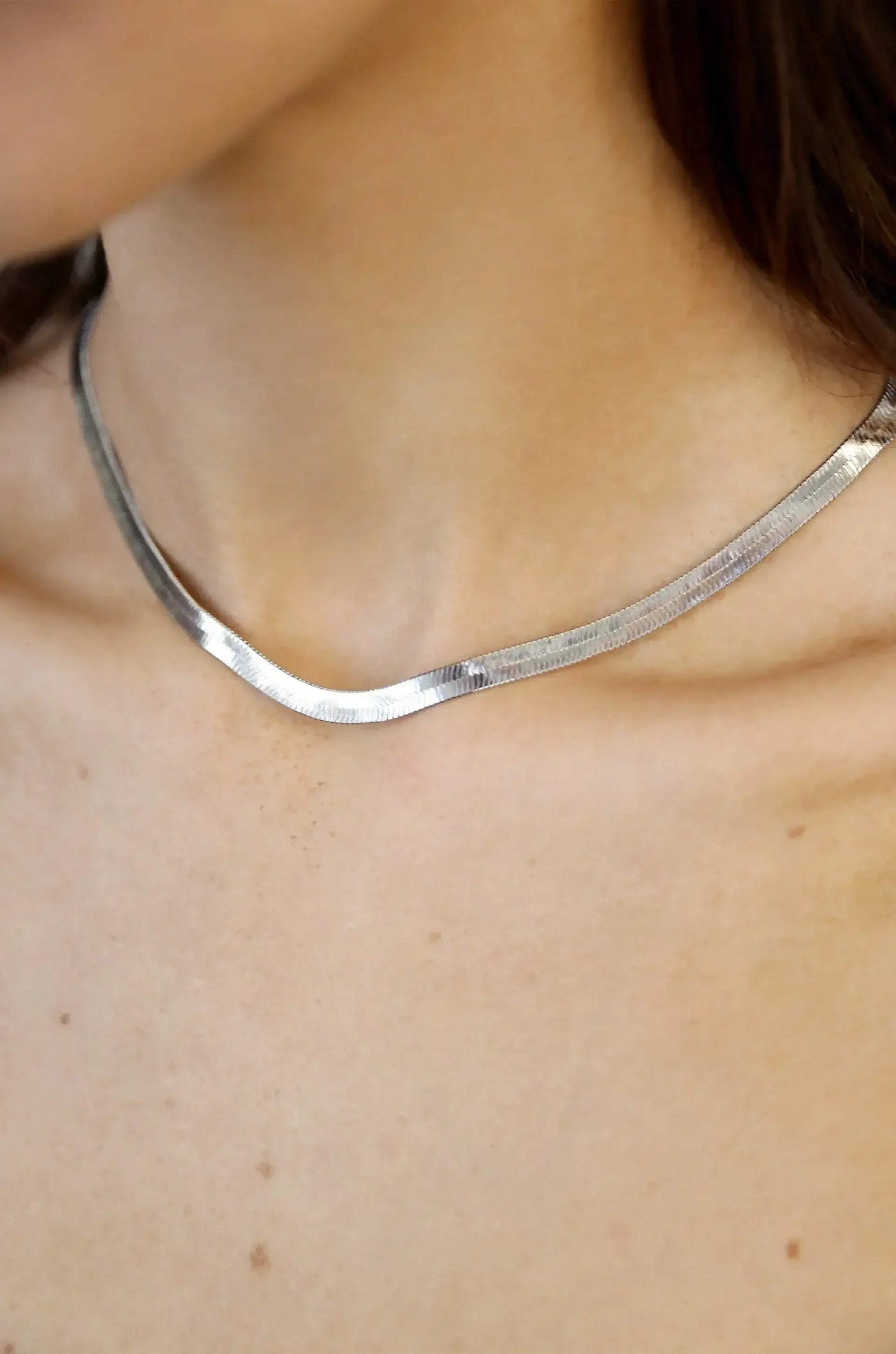 A silver necklace rests elegantly around a personâ€™s neck, reflecting light against their smooth skin, which features subtle freckles in a neutral-toned setting.