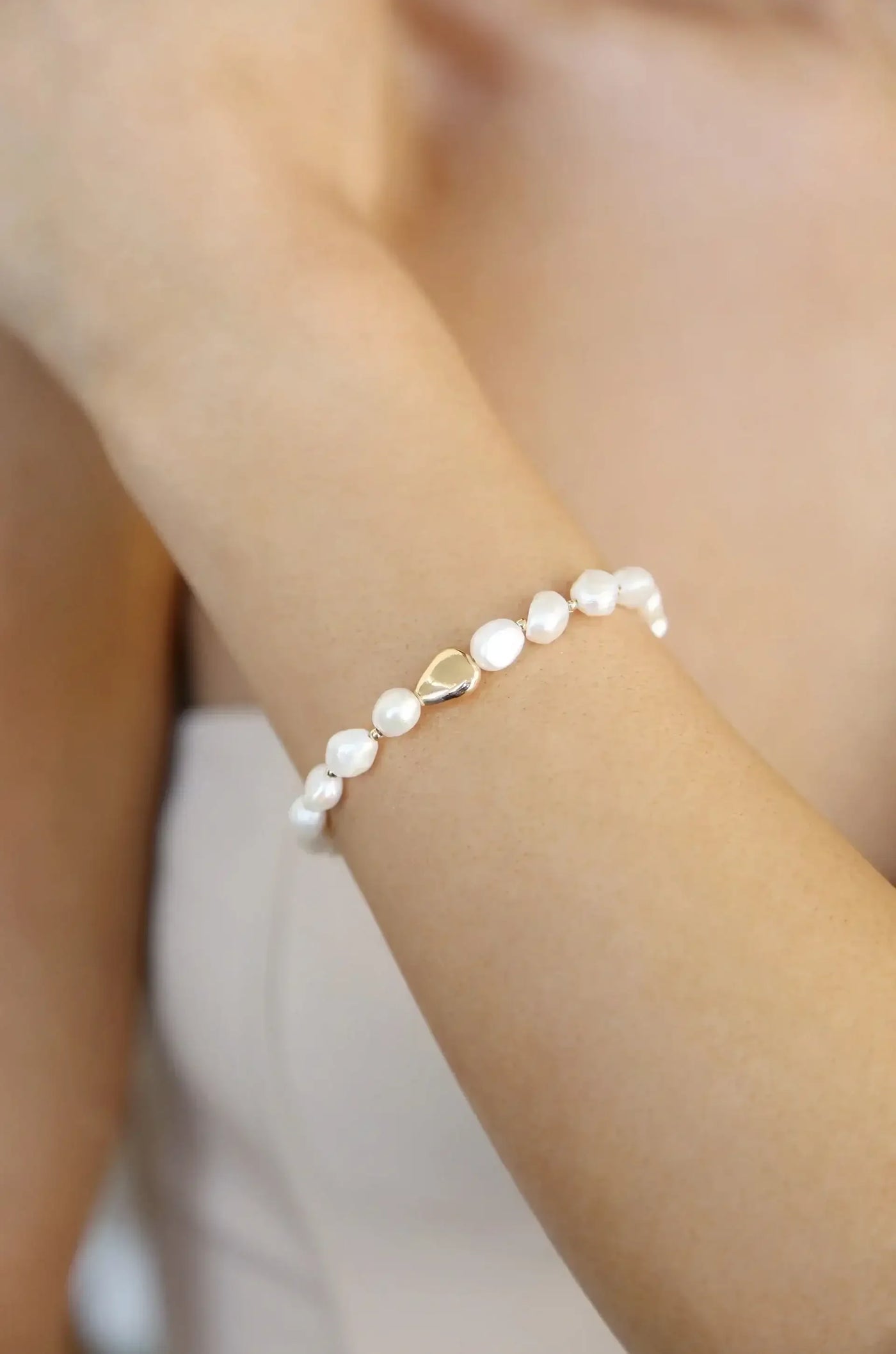 A delicate bracelet made of irregular pearls with a teardrop-shaped gold clasp adorns a tanned arm, conveying elegance in a neutral setting.