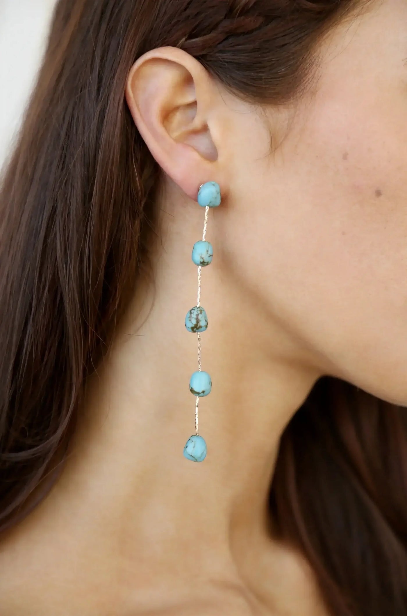 A pair of long, dangling earrings featuring several turquoise beads is worn on the left ear of a young woman, highlighting her skin tone and long, brown hair.