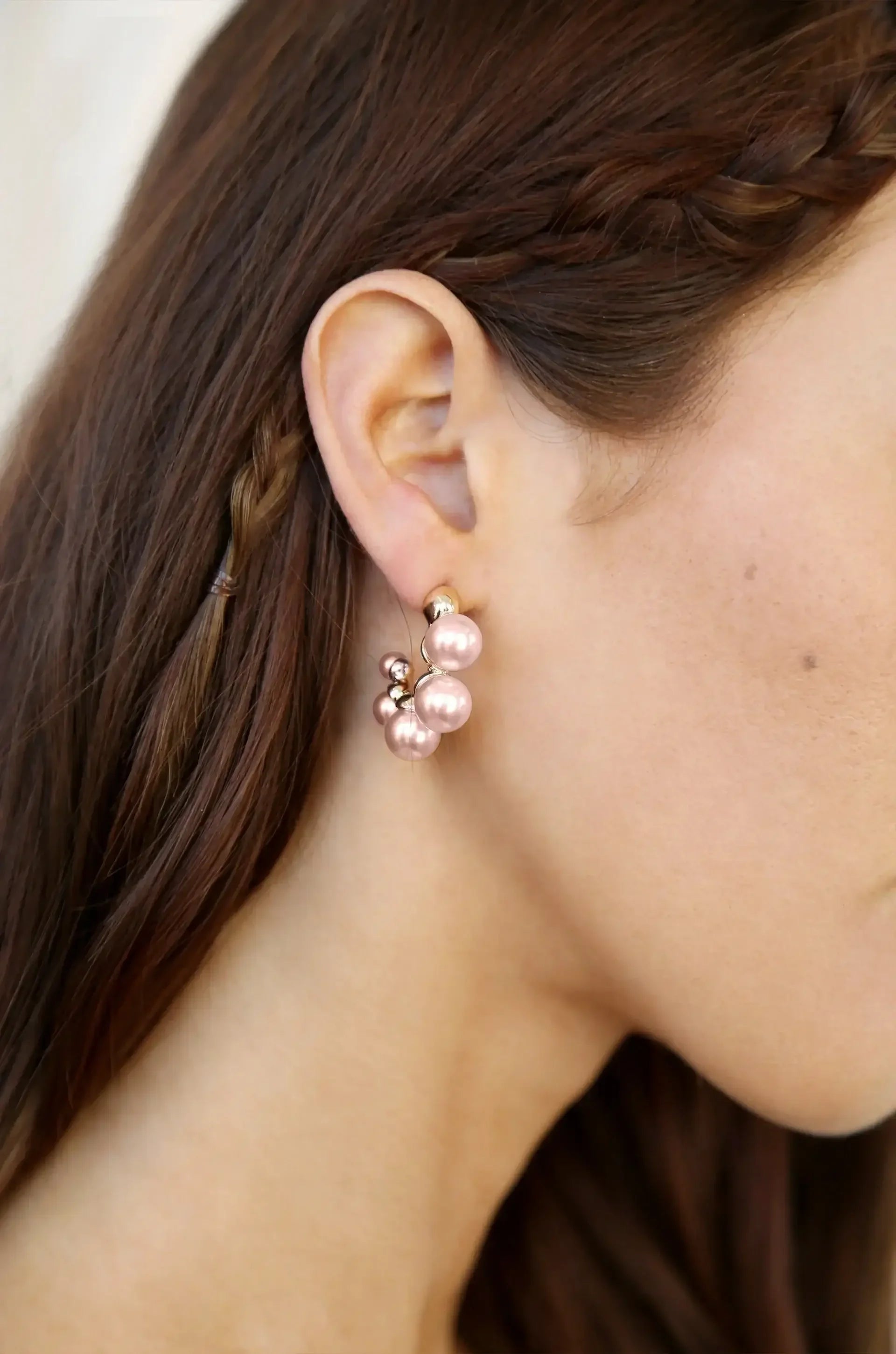 A close-up of a woman's ear adorned with pink pearl earrings. The earrings feature a cluster design and are set against a backdrop of smooth, dark brown hair, partially braided.