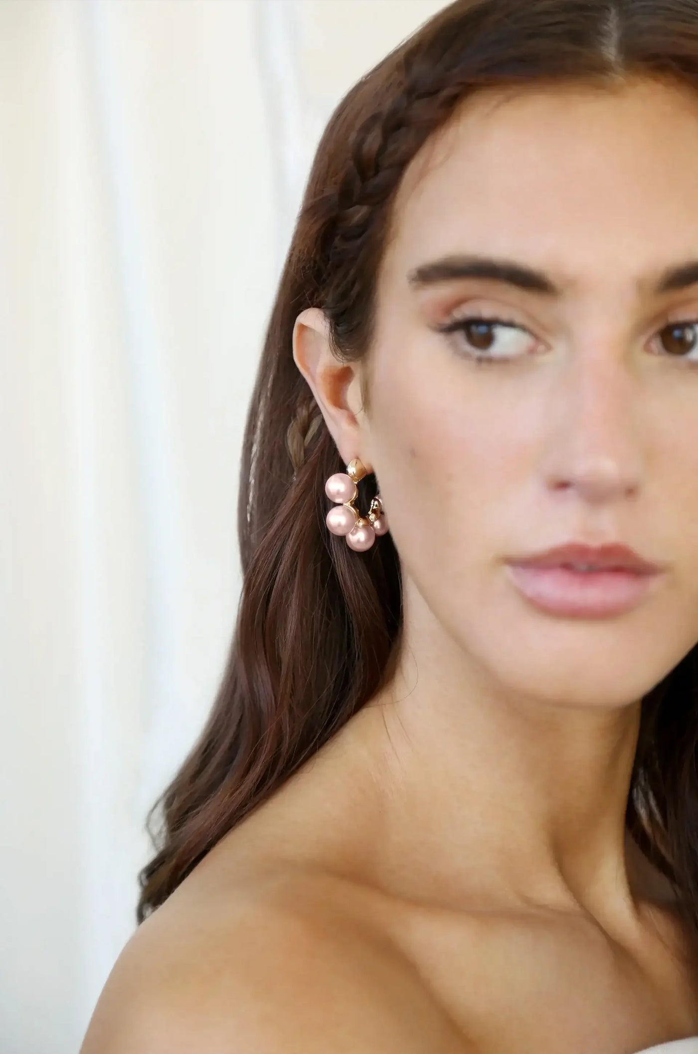 A young woman with long, dark hair and subtle makeup showcases a large, pink pearl earring. She appears in a softly lit, neutral-toned background, highlighting her features and jewelry.