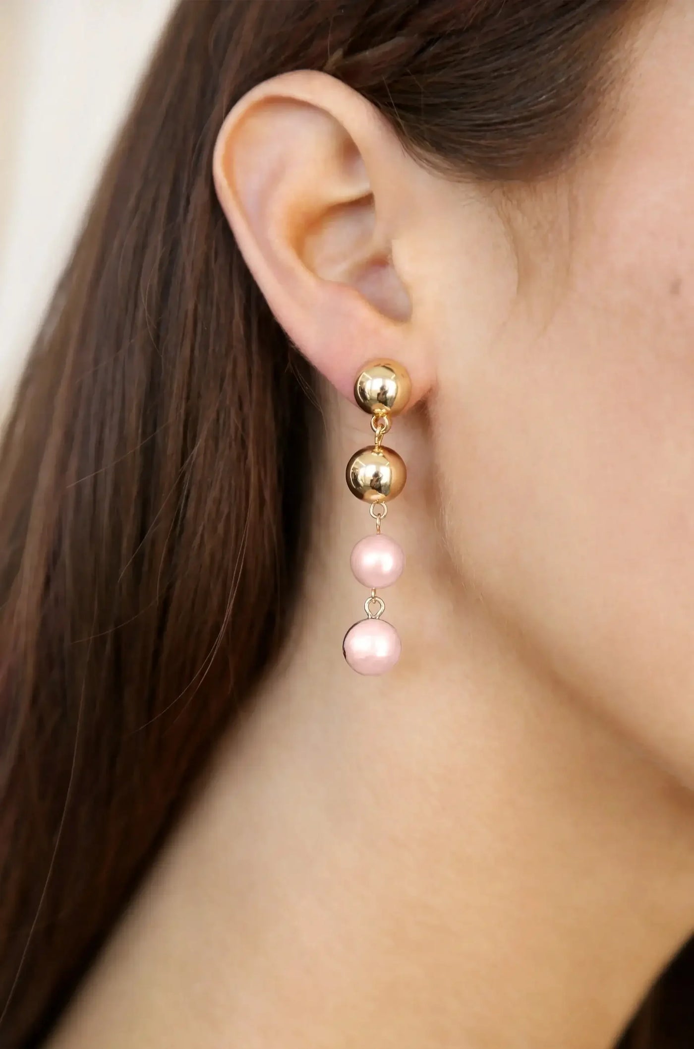 A pair of earrings features a gold stud at the top, dangling two pink pearls below. The earrings are adorned on the ear of a person with long brown hair, set against a neutral background.