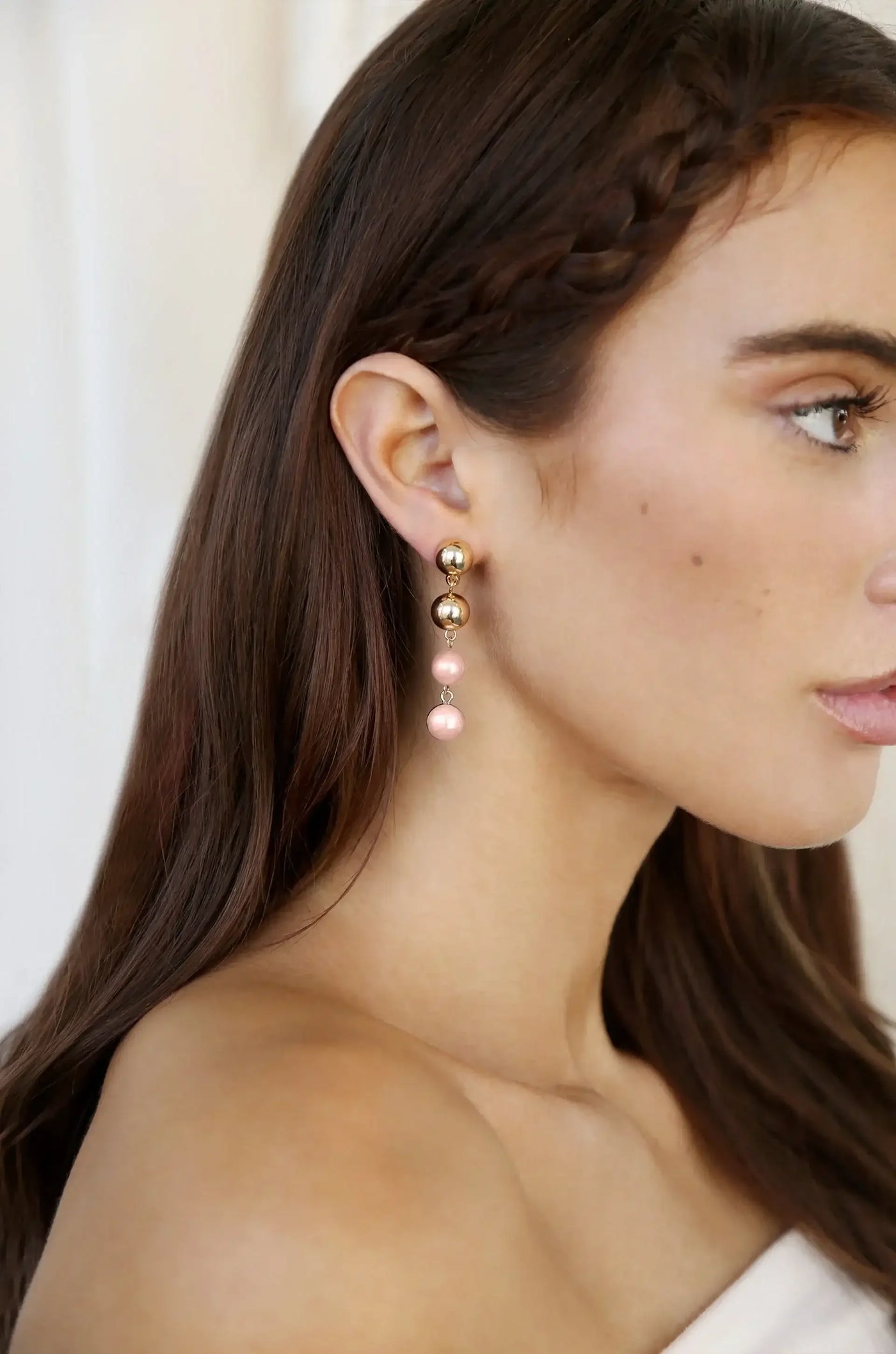 A woman with long, dark hair wears elegant drop earrings featuring gold and pink spheres. She poses gently to the side, showcasing her accessories against a soft, blurred background.