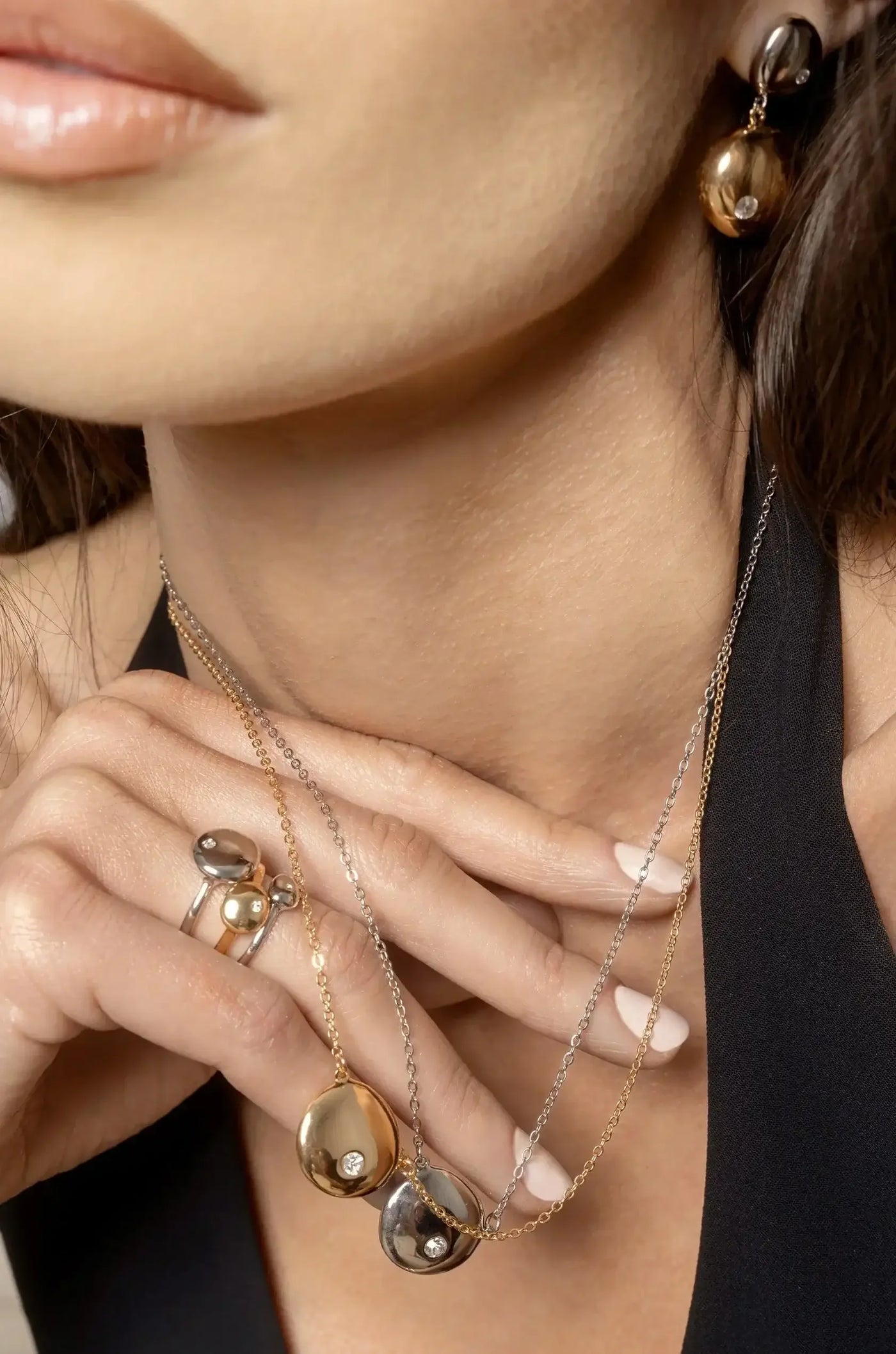 A close-up of a woman's hand adorned with rings, gently holding necklaces featuring metallic and crystal elements, against a backdrop of her neckline. The skin is smooth and lightly illuminated.