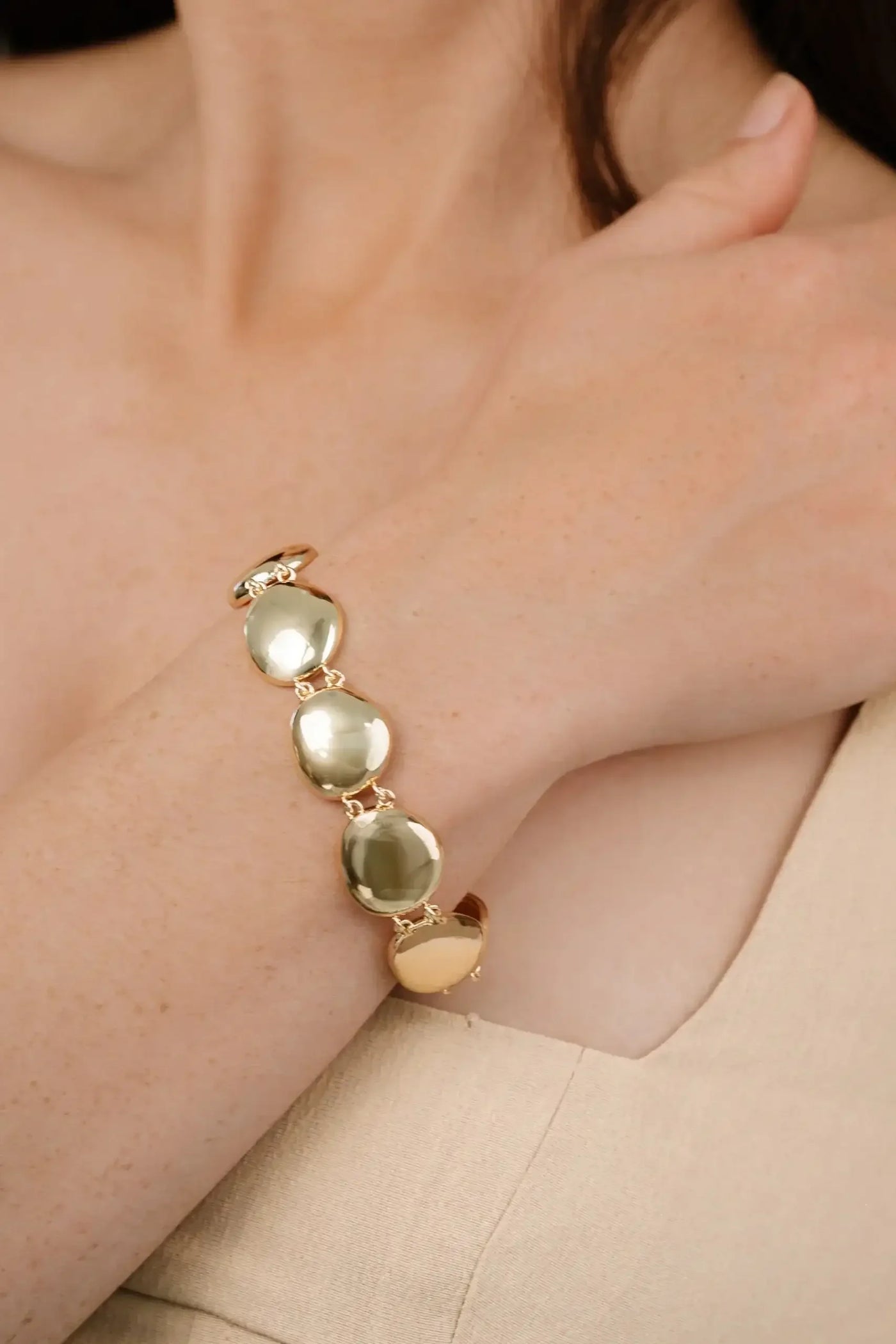 A gold bracelet with three round, reflective stones is worn on a wrist. The hand gently rests against the arm, set against a soft, neutral background.