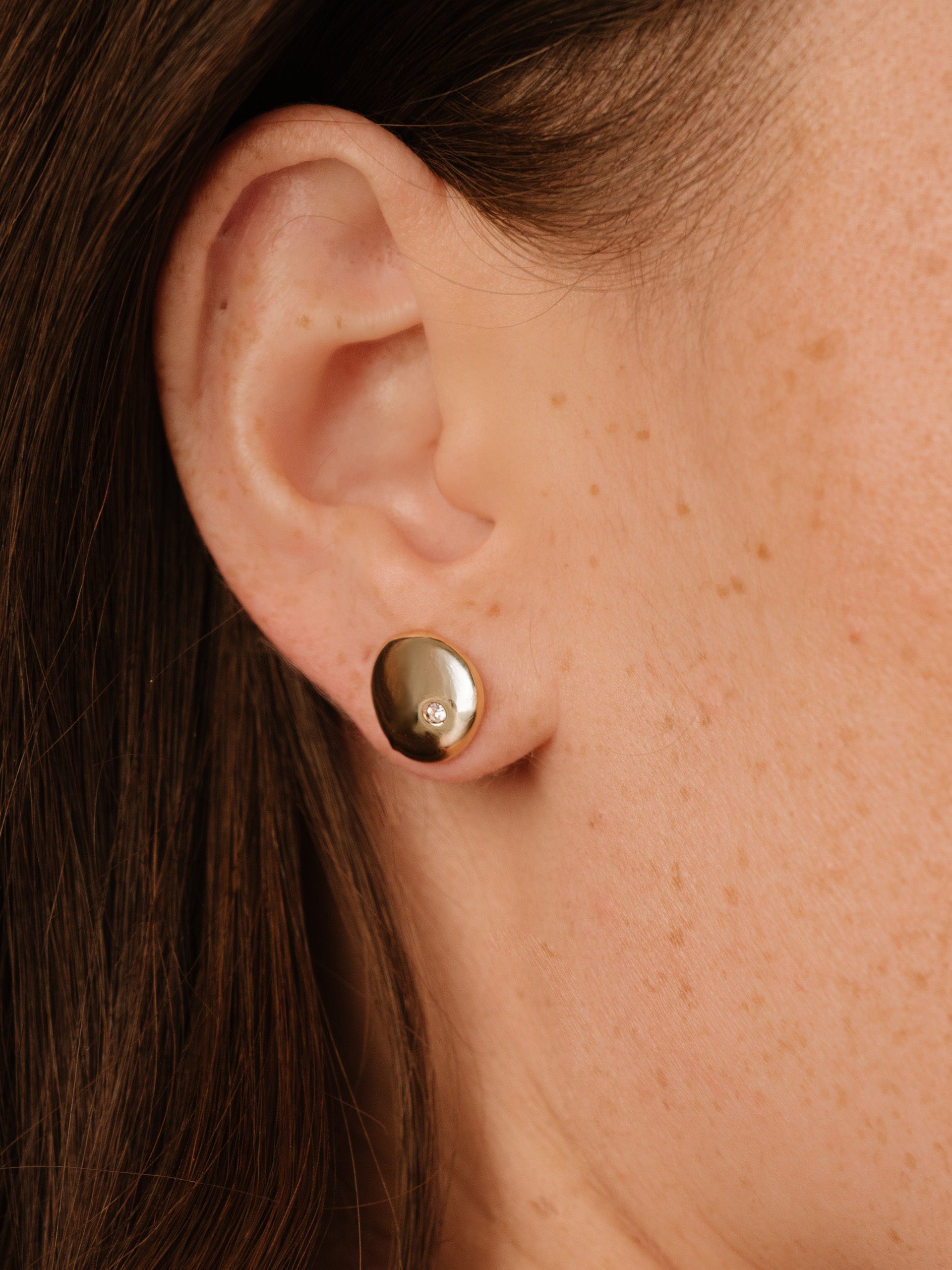 A gold earring is prominently displayed on the ear of a person, featuring a small gemstone. The setting is close-up, highlighting the texture of the skin and earring.