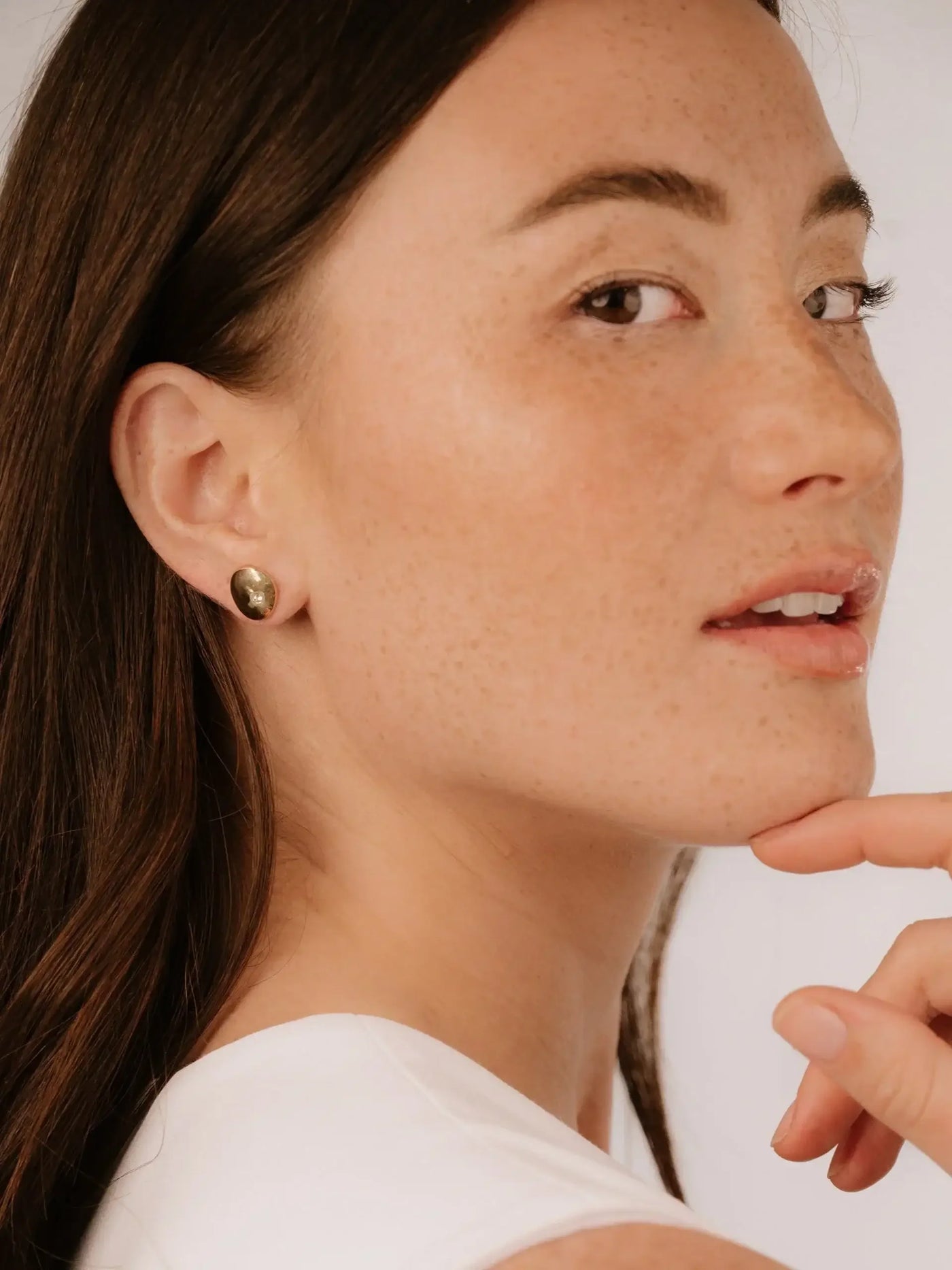 A woman with long brown hair and freckles gently touches her chin while looking slightly over her shoulder, wearing a simple white top and stud earrings against a light background.