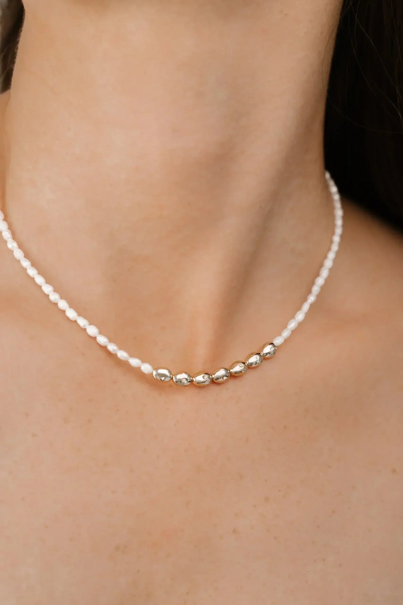 A delicate necklace rests on a person's neck, featuring alternating white beads and gold oval accents, against a background of smooth skin and dark hair.