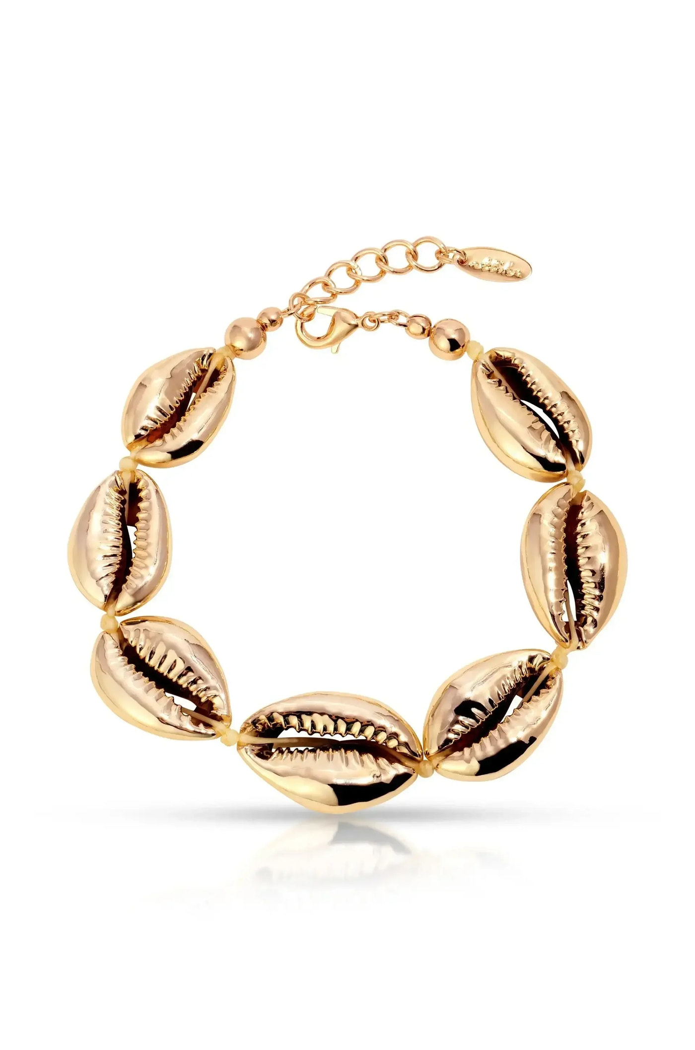 A gold bracelet features a series of shiny cowrie shell motifs, connected by small beads, with an adjustable chain for size. It rests on a reflective surface.