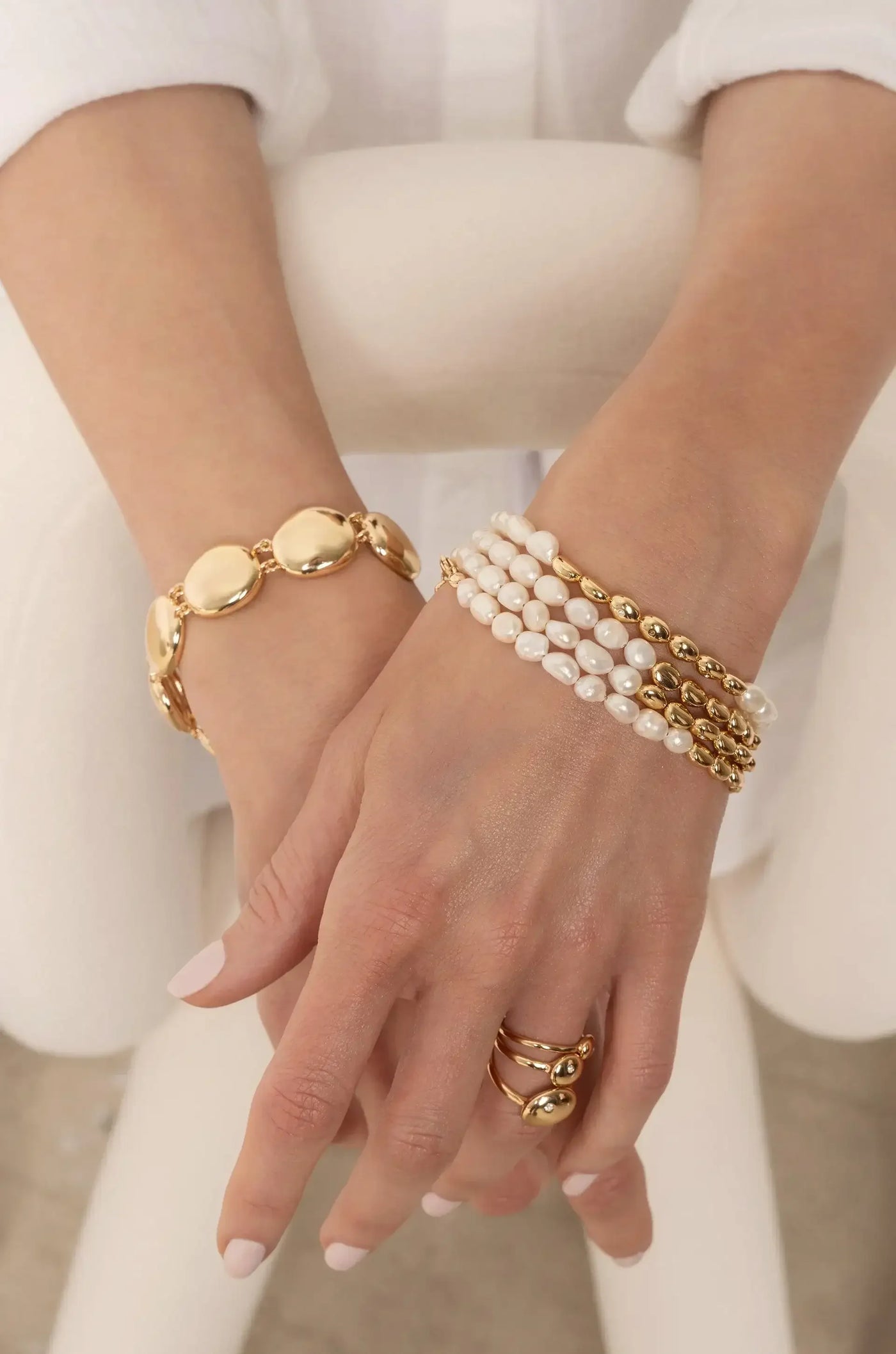 Two elegantly adorned hands display a gold bracelet and multiple pearl and gold bead bracelets, arranged harmoniously against a soft, neutral background that enhances the jewelry's shine.