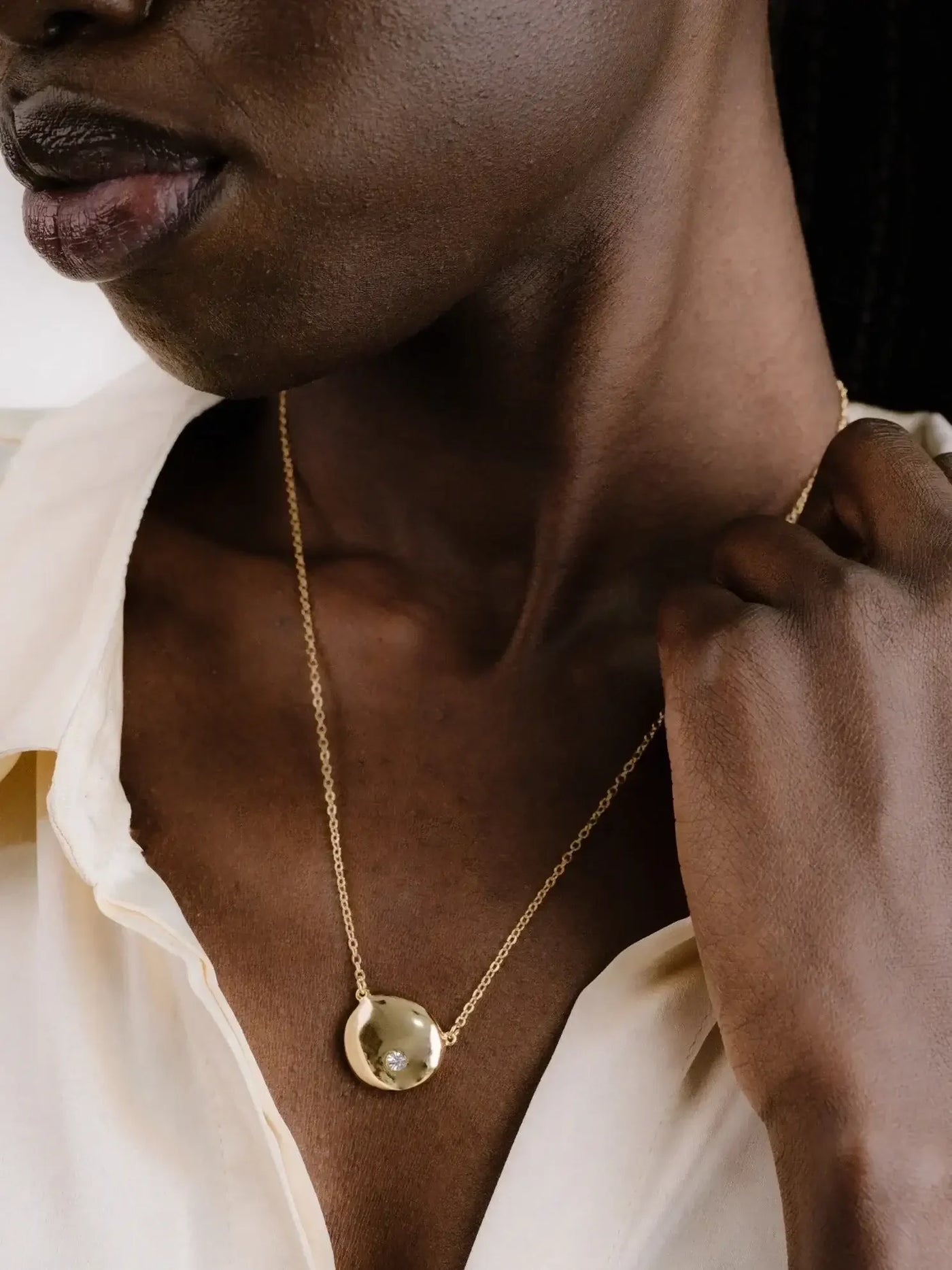 A close-up of a person's neck and shoulder, adorned with a gold necklace featuring a round pendant, highlights smooth skin and a subtle, elegant expression. The background is softly blurred.