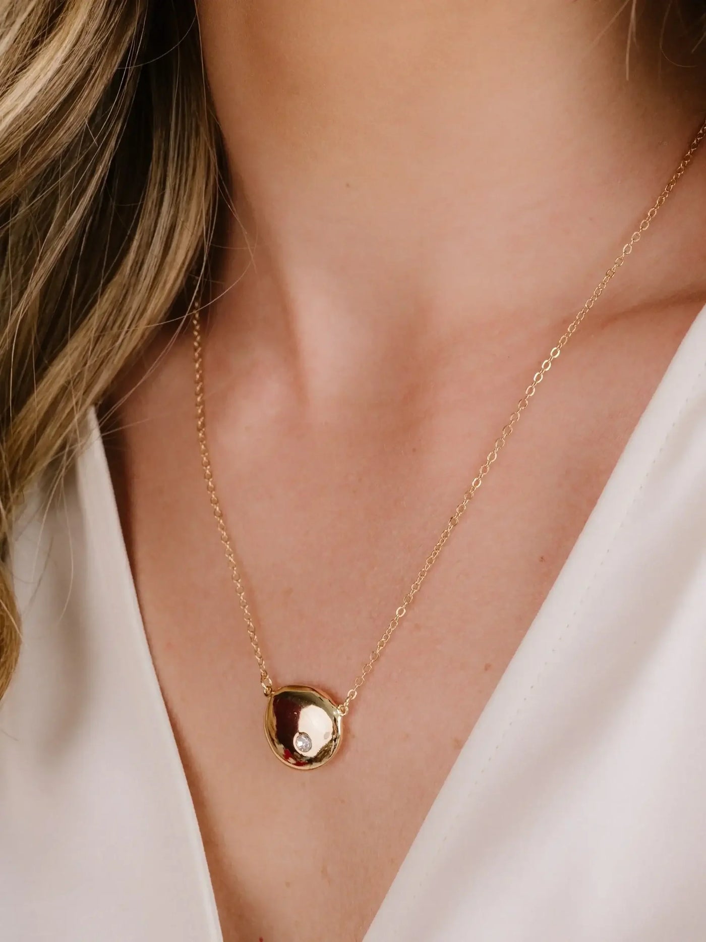 A gold necklace with a round pendant featuring a small gem is resting on the collarbone of a person, who is wearing a white top. The background is softly blurred.