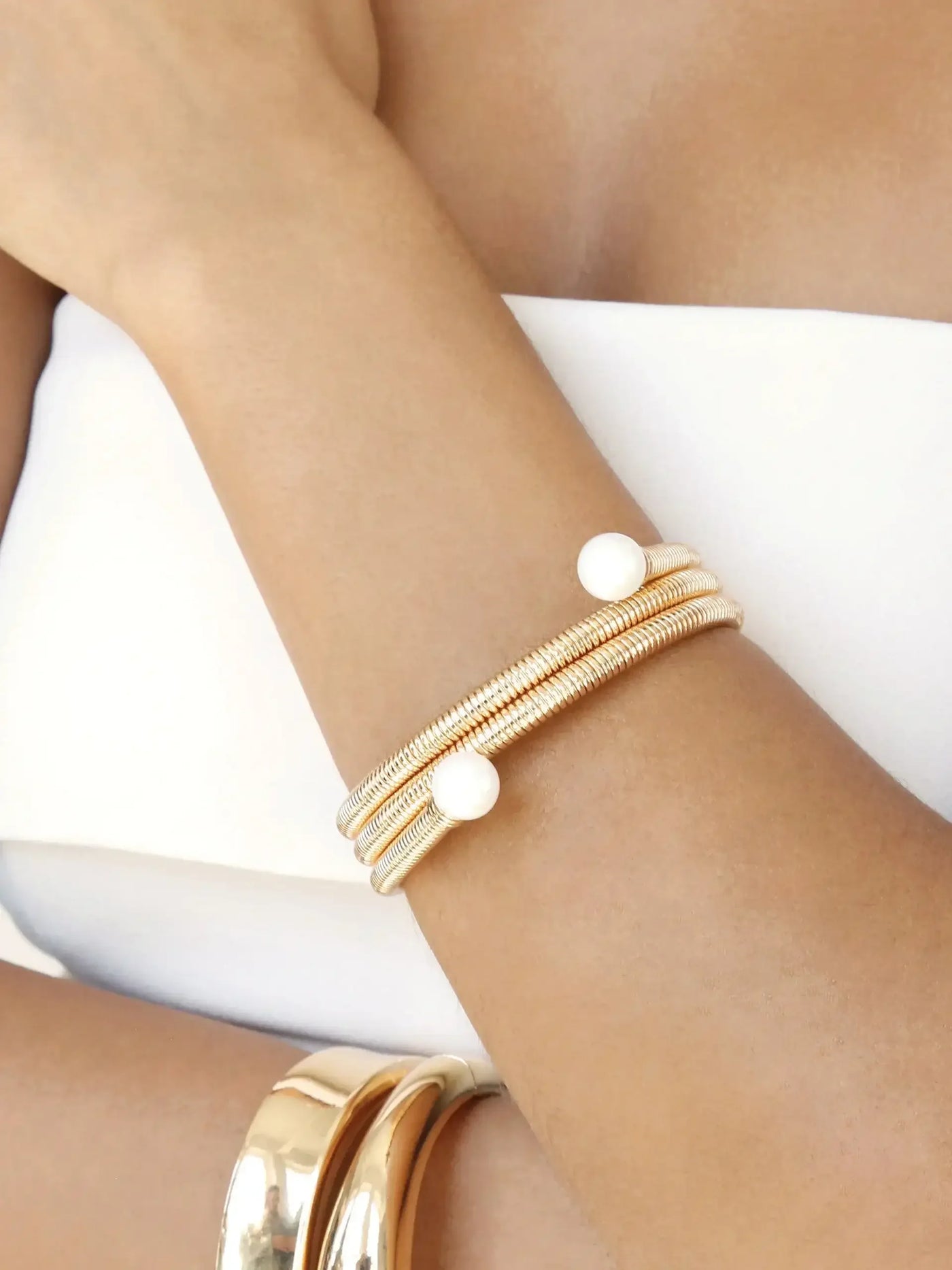 A person's wrist adorned with multiple gold bracelets, including one with a white pearl, rests against a light-colored background. The arm appears relaxed and well-groomed.