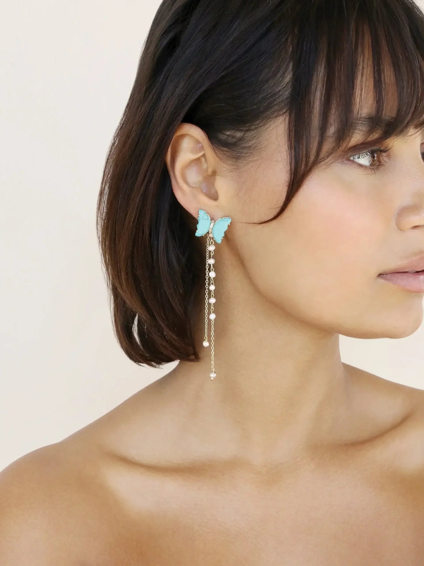 A woman is wearing turquoise butterfly earrings that have a cascading chain of small beads, set against a neutral background, highlighting her shoulder-length hair and bare shoulders.