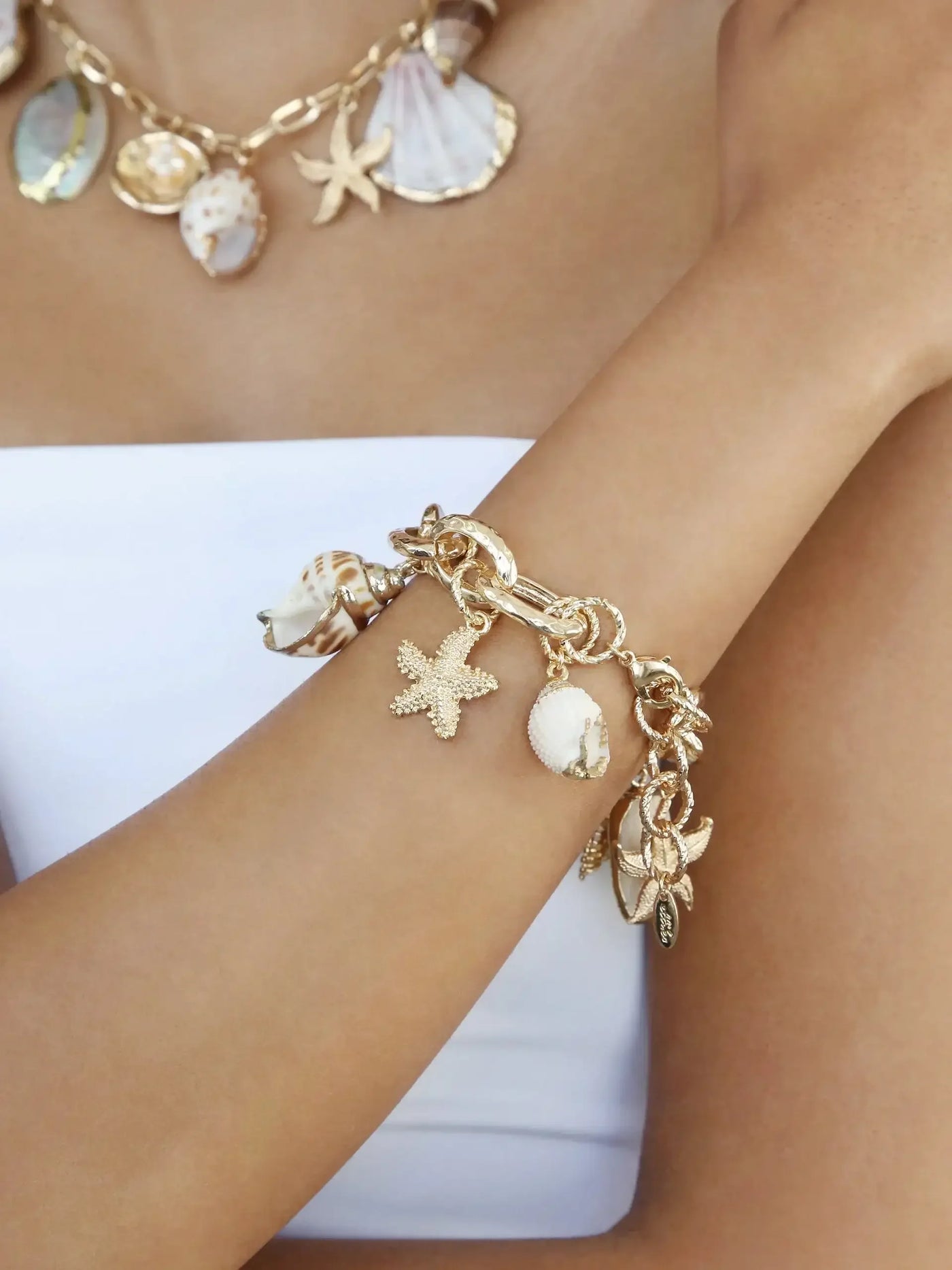 A gold charm bracelet adorned with seashells and starfish hangs on a tanned arm. The arm is positioned against a white background, complementing the beach-themed jewelry. A shell necklace is visible nearby.