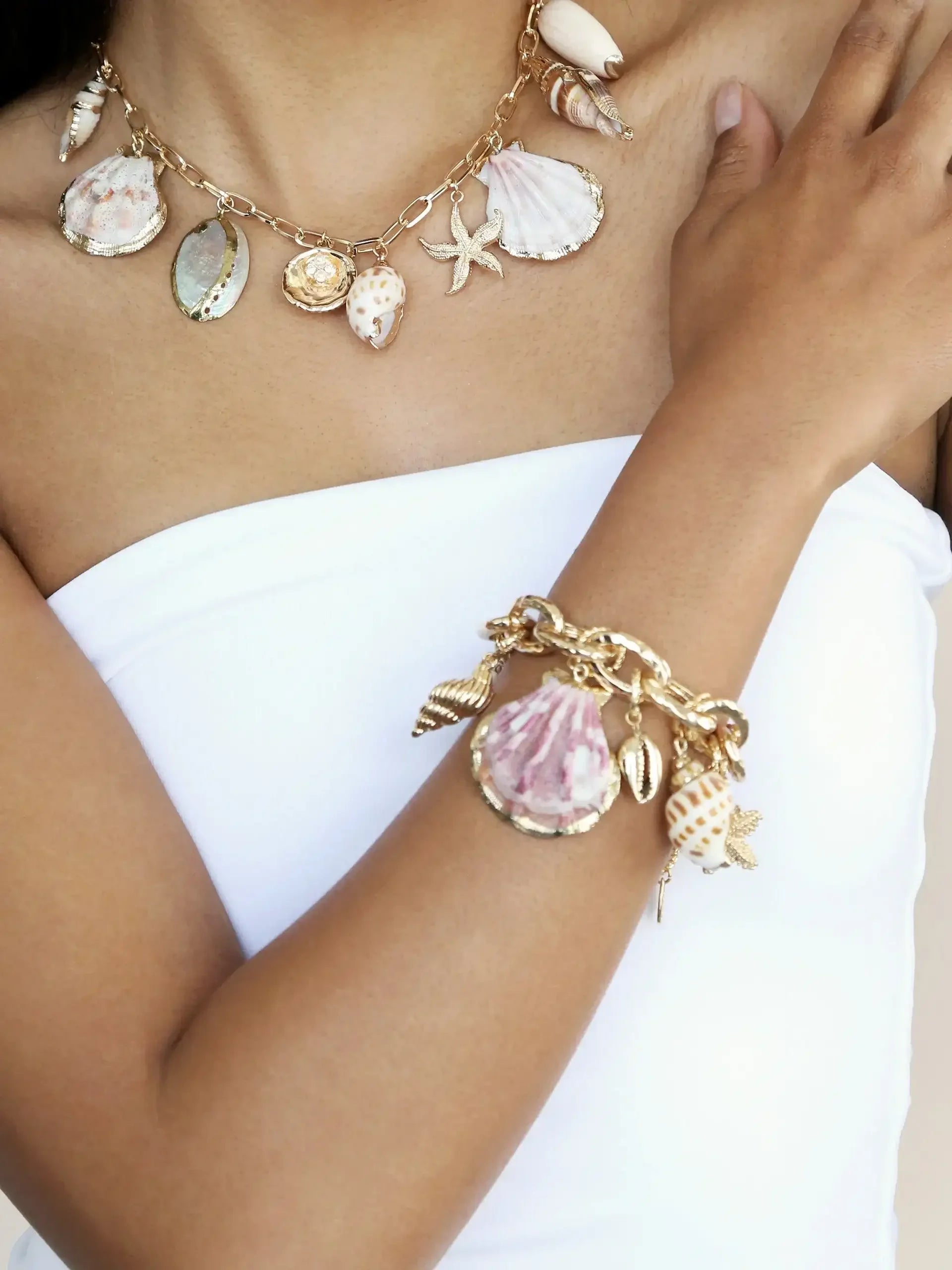 A necklace and bracelet adorned with various seashells and a starfish drape elegantly over a bare shoulder, creating a beach-like aesthetic against a neutral background.