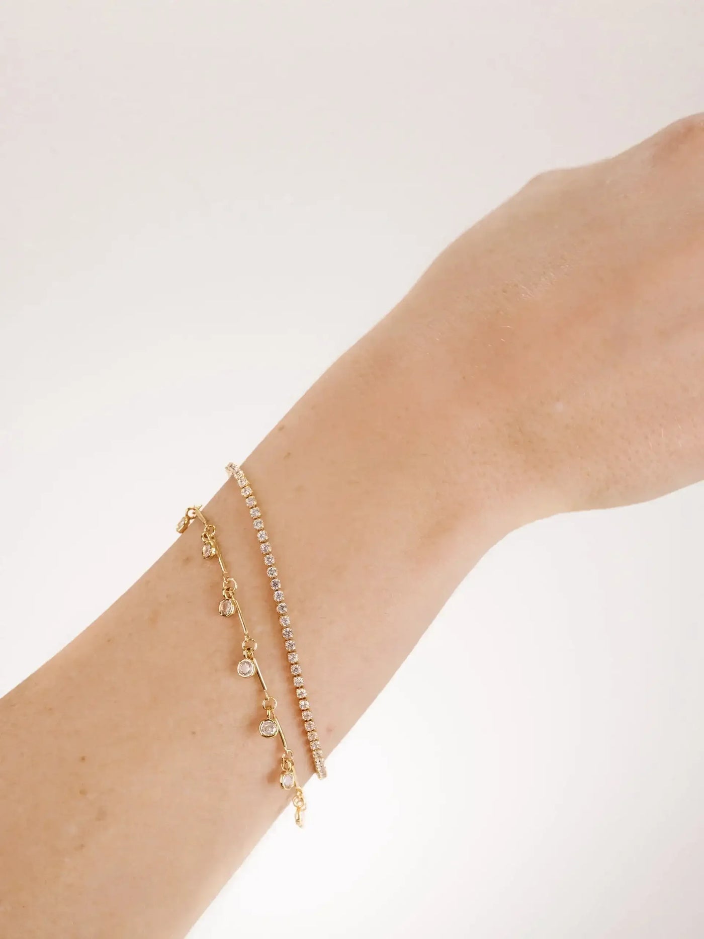 A hand with a slender wrist showcases two gold bracelets: one studded with sparkling stones and another with dangling gems. The background is a soft, light color.