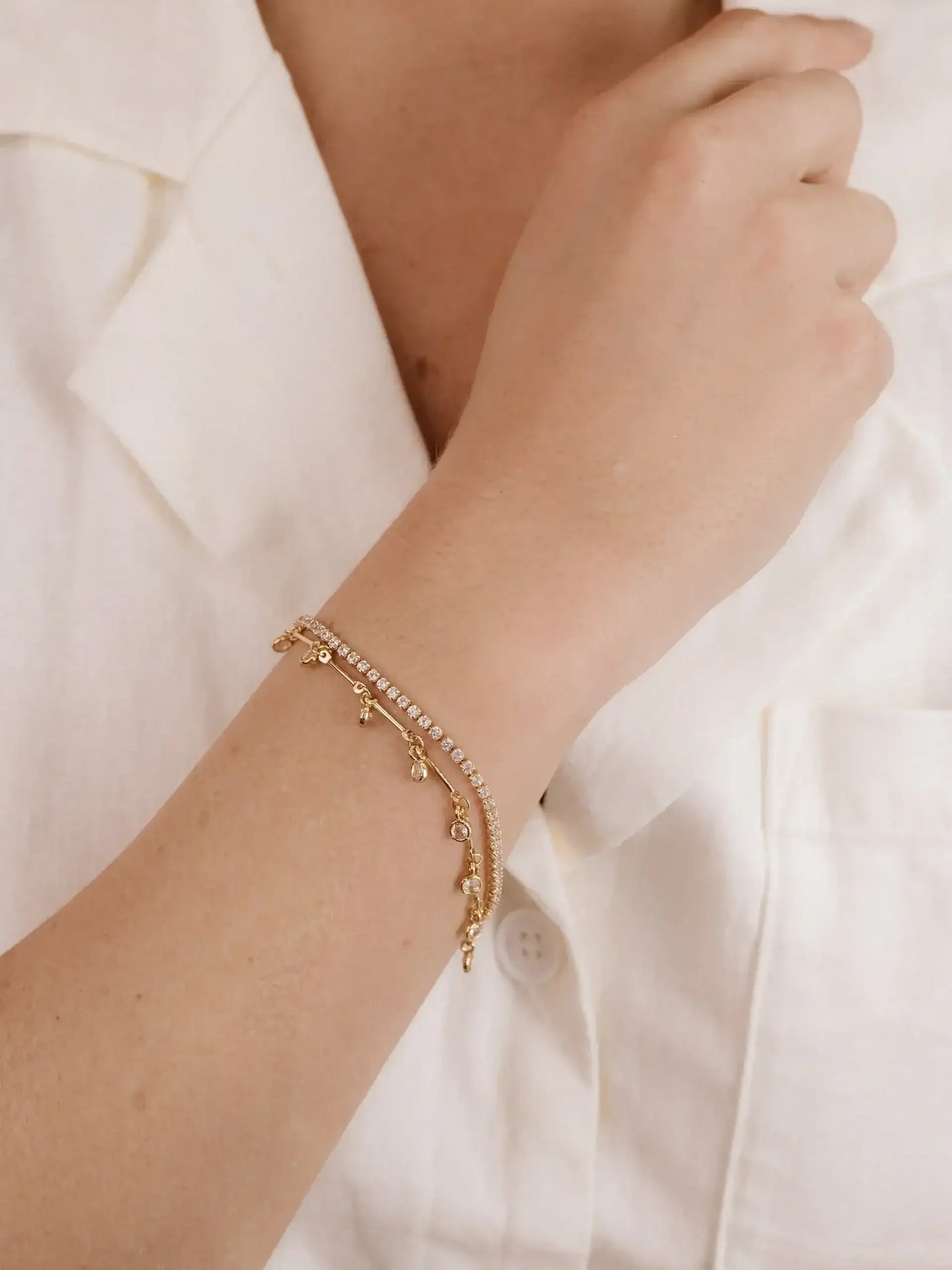 A gold bracelet adorned with small accents rests on a wrist, enhancing the arm's elegance. The wearer is dressed in a light-colored, buttoned shirt, suggesting a casual yet refined setting.