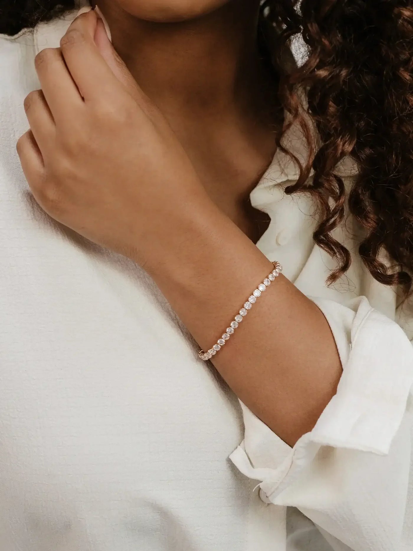 A hand with a light brown skin tone is gently touching a collarbone, adorned with a delicate, shimmering bracelet of small, circular stones. The person wears a white collared shirt.