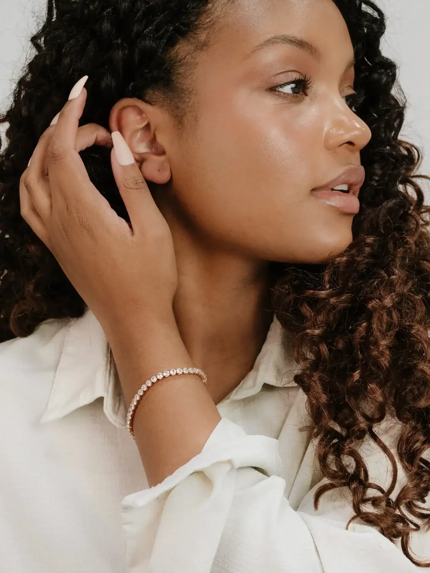 A woman with curly hair gently touches her ear with one hand while the other rests on her arm, adorned with a sparkling bracelet, set against a neutral background.