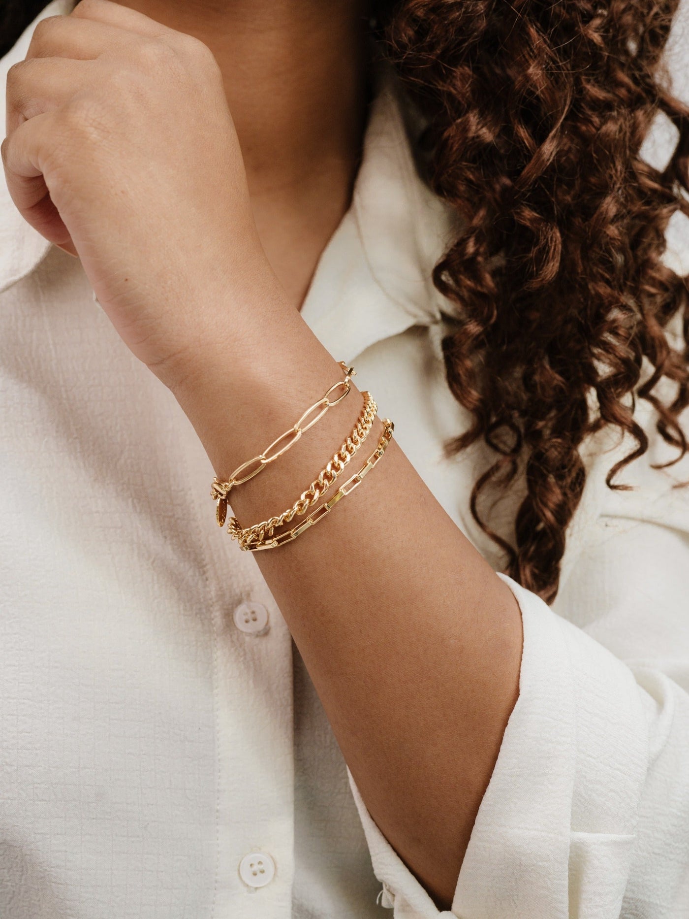 A hand adorned with multiple gold bracelets rests against a textured, white shirt. The bracelets feature different patterns and styles, enhancing the elegant appearance of the arm.