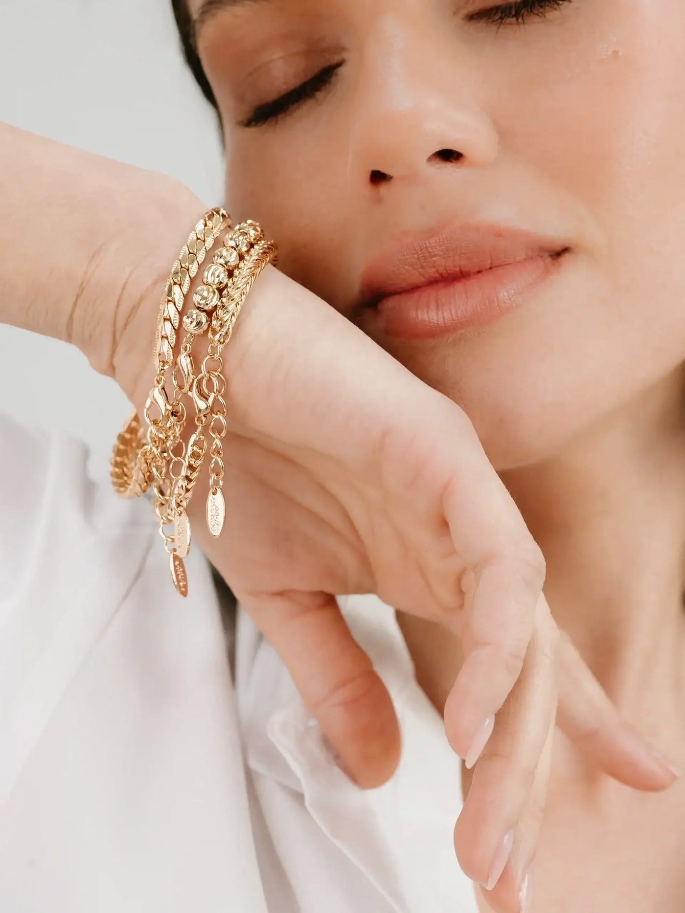 A person's hand, adorned with multiple gold bracelets, rests near their face. The individual appears relaxed, with eyes closed and lips slightly pursed, set against a neutral background.