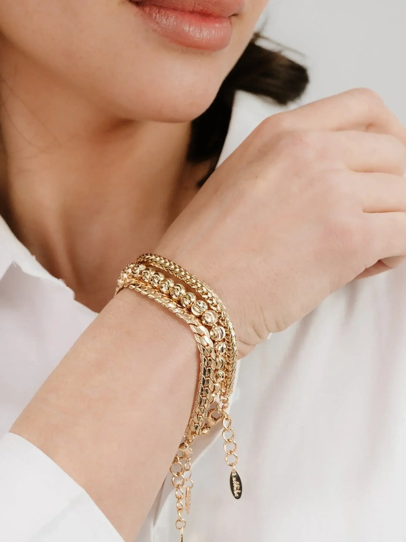 A hand adorned with multiple gold bracelets rests against a light-colored, textured background, showcasing a stylish accessory against the skin. The person wears a white shirt, highlighting the jewelry.