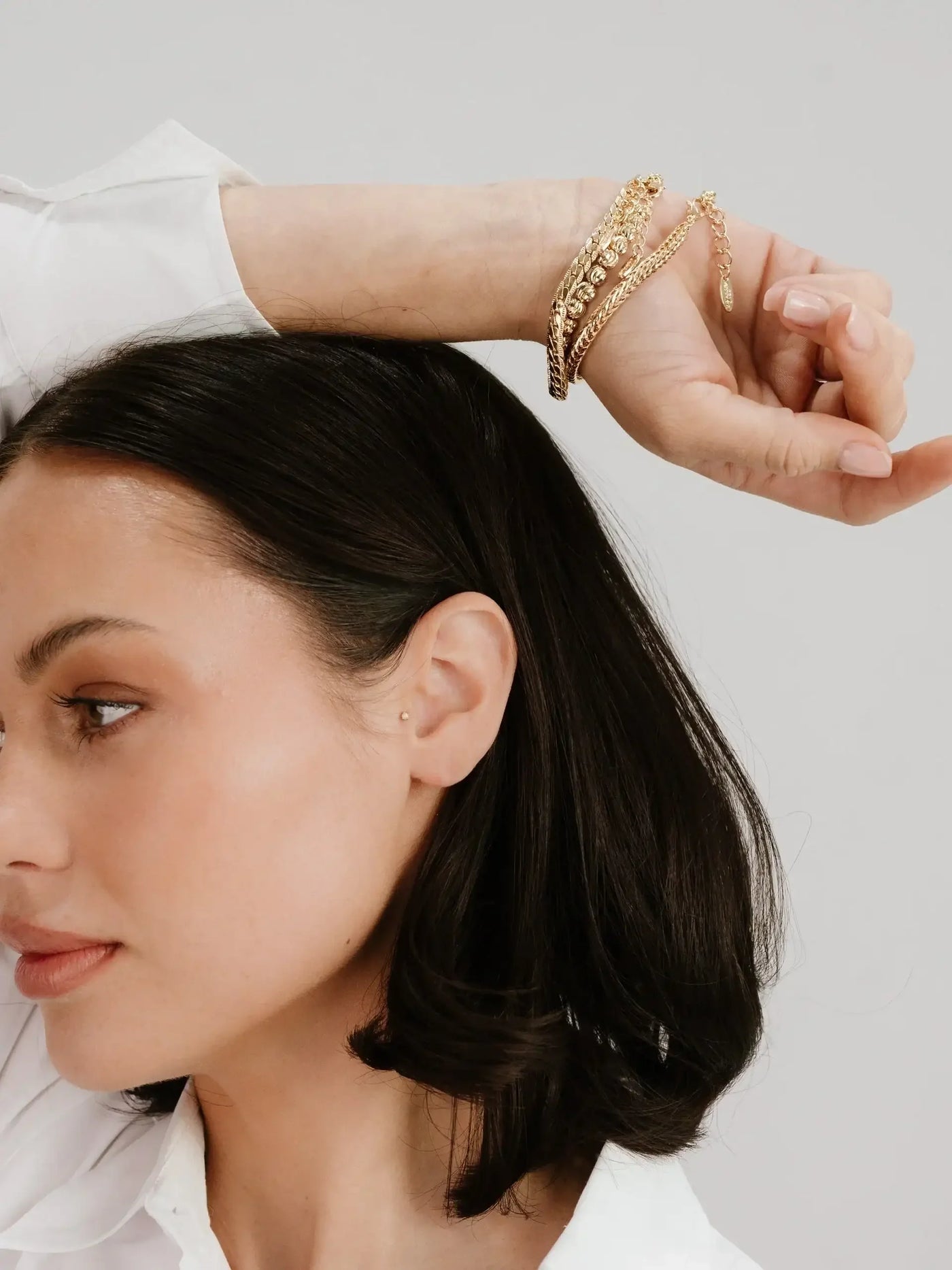 A woman with dark hair is holding her wrist, adorned with multiple gold bracelets. She gazes to the side against a light gray background, wearing a white shirt.