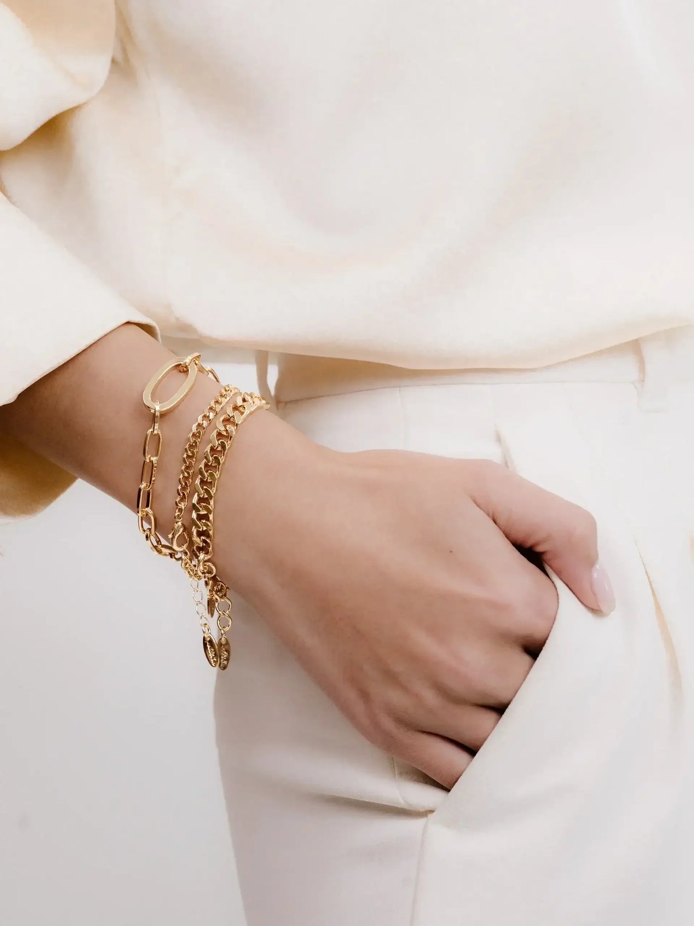A hand adorned with multiple golden bracelets rests casually in the pocket of pale, tailored pants, suggesting a relaxed yet stylish demeanor in a soft, light-colored environment.