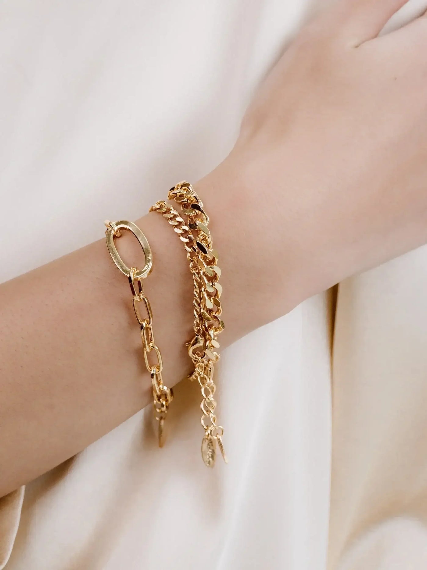 A hand adorned with three gold bracelets rests against a softly draped, light-colored fabric. The bracelets vary in design, featuring links and charms, reflecting a stylish accessory choice.