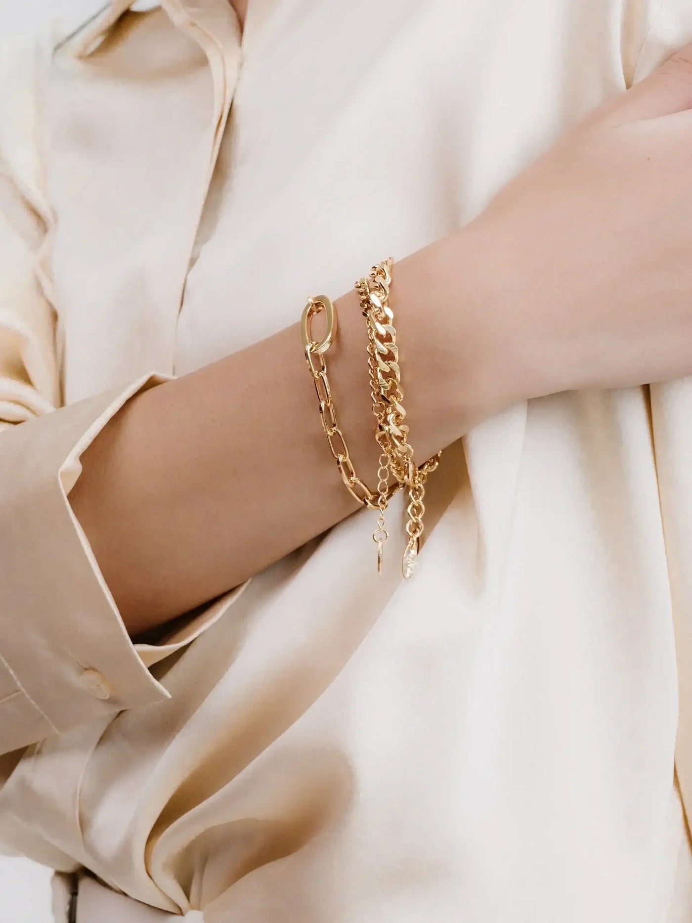 A hand adorned with multiple gold bracelets rests against a light, silky fabric. The bracelets feature varying chains and designs, showcasing a blend of elegance and style.