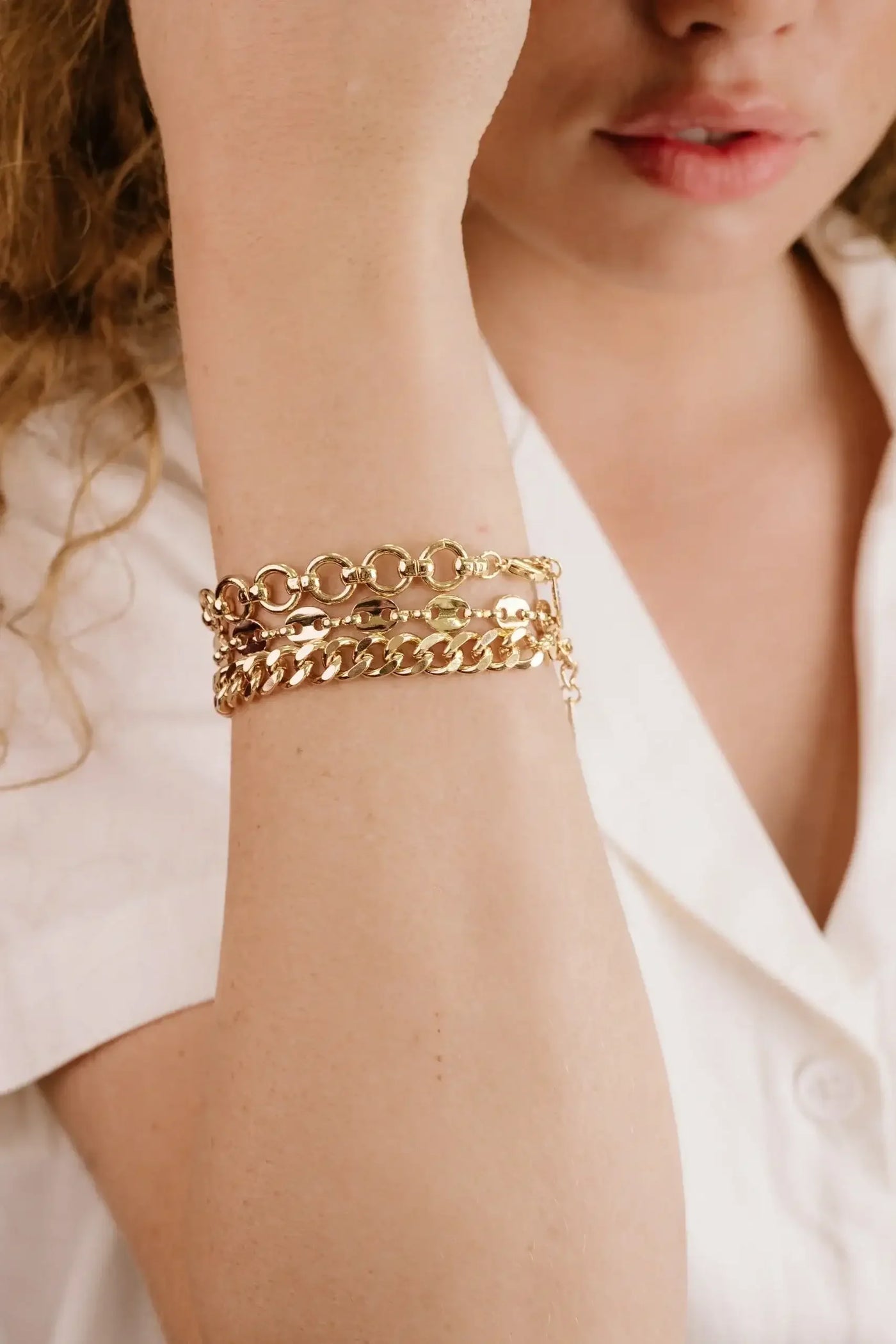 A womanâ€™s arm showcases multiple gold bracelets, with one hand raised to her face. She wears a white shirt, and her lips are slightly parted, suggesting a contemplative expression.