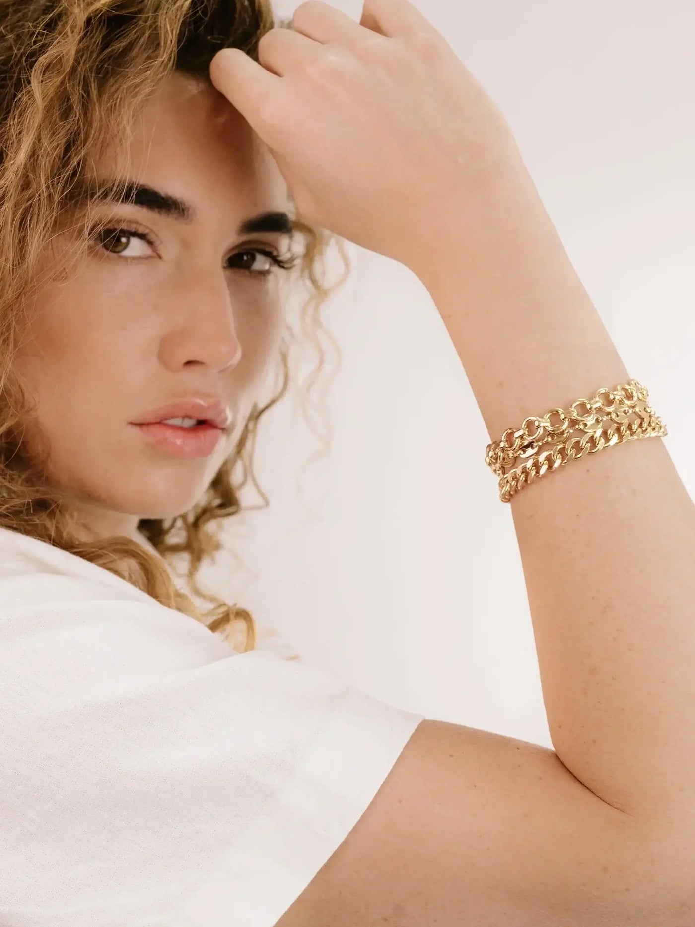 A woman with wavy, curly hair poses while touching her hair, wearing a stylish gold chain bracelet on her wrist, set against a light, neutral background.