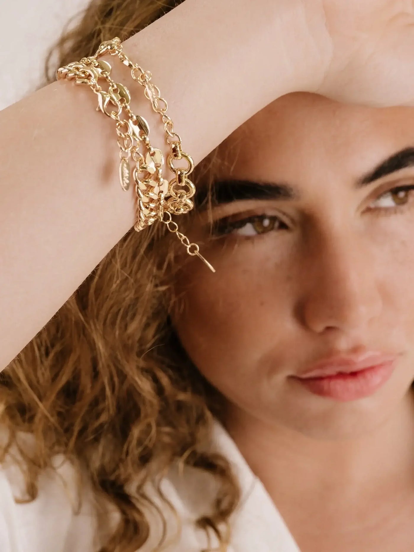 A gold chain bracelet adorns a womanâ€™s wrist as she gently rests her arm against her forehead. Her curly hair and natural makeup complement the elegant jewelry, set against a neutral background.