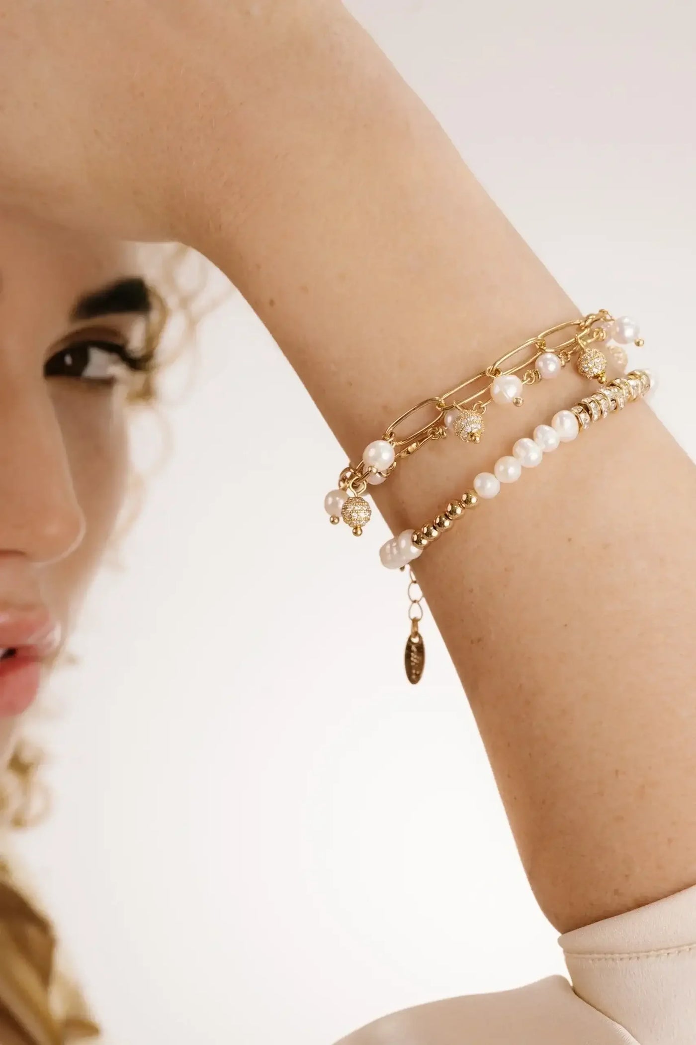 A hand adorned with multiple elegant bracelets is raised near the face of a model, who has curly hair and appears to be posing against a soft, blurred background.